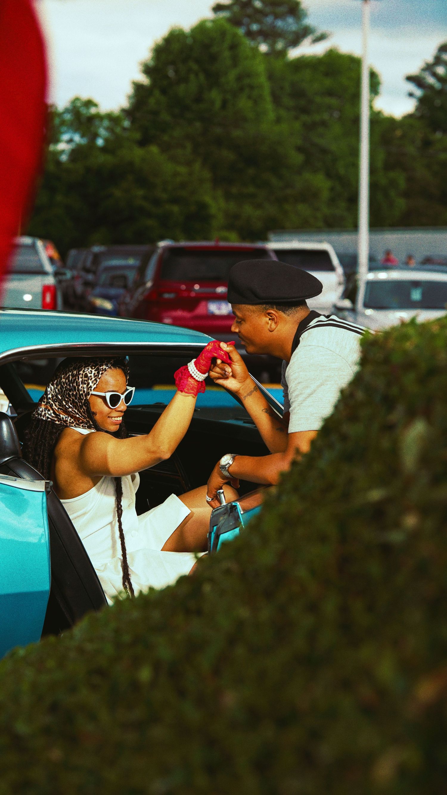 Two people sharing a playful moment through a teal classic car window in a parking lot.