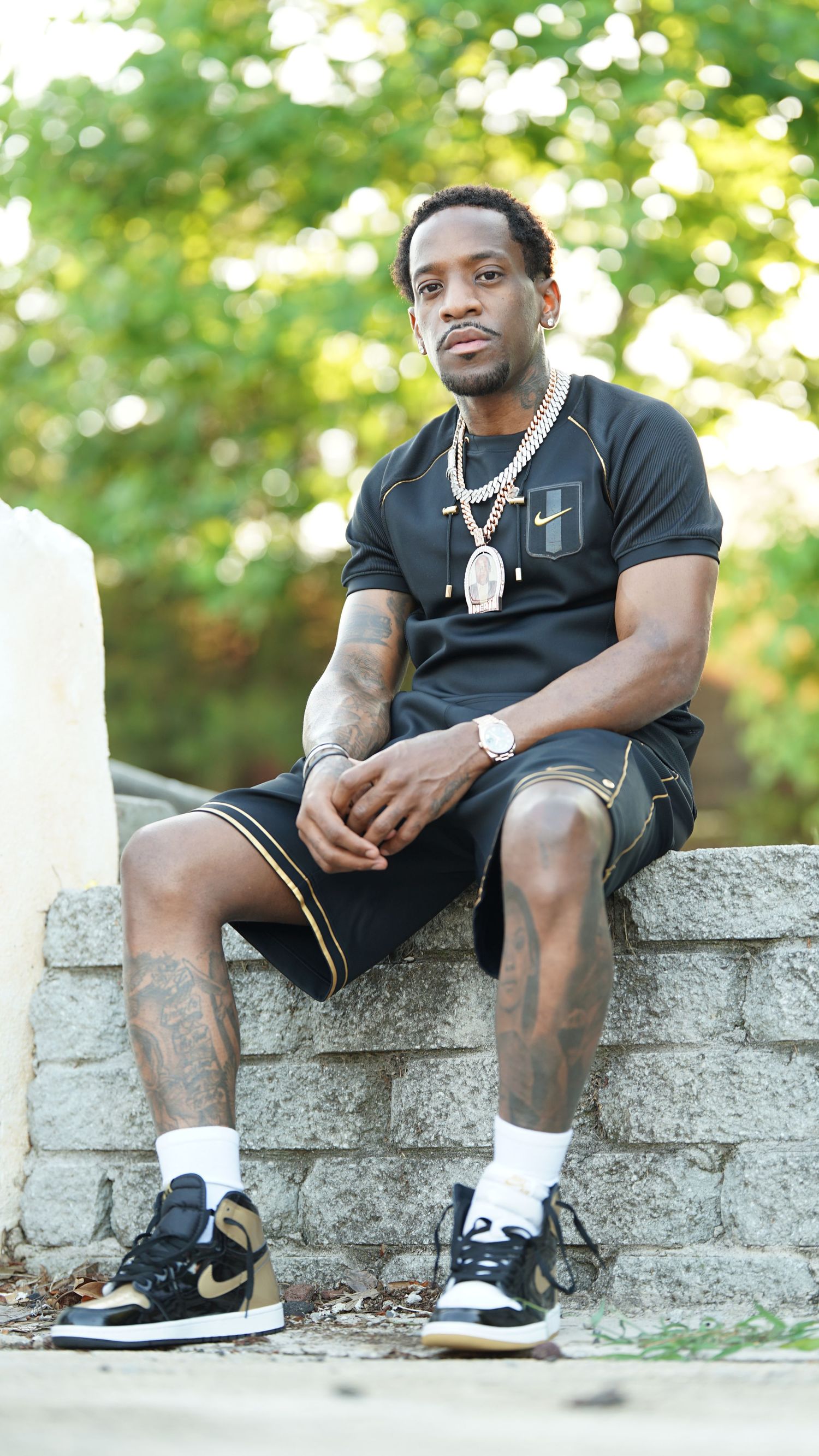 A person in black clothing sits casually on a concrete wall outdoors wearing Nike sneakers and jewelry.