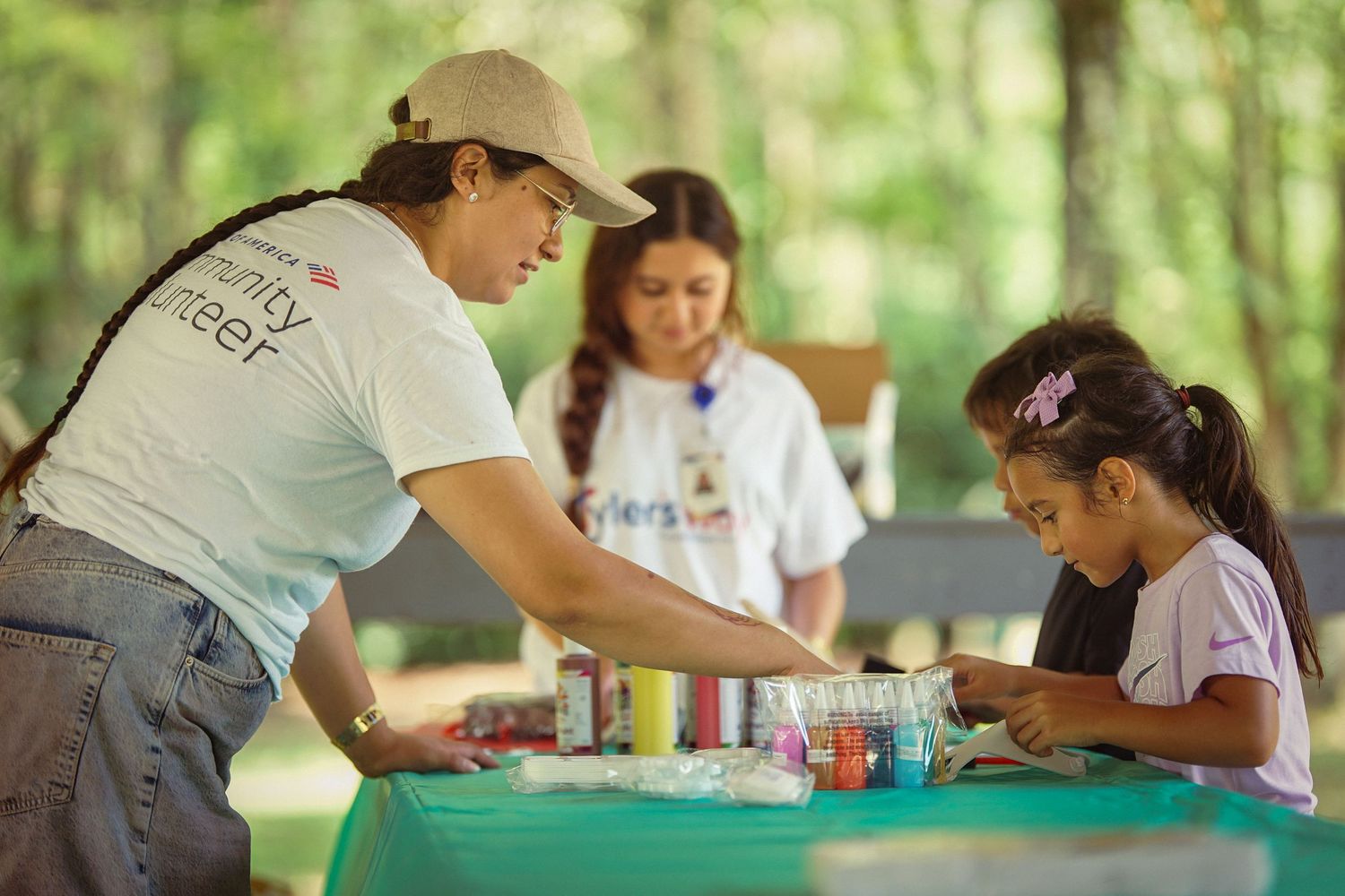 A volunteer in a cap helps a young girl with a craft activity at an outdoor community event table.