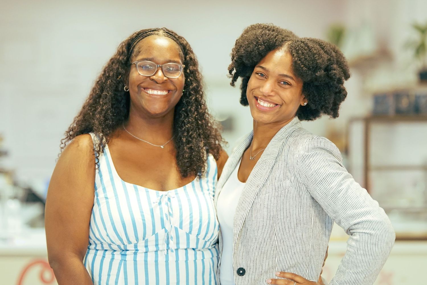 Two smiling women posing together in a bright indoor setting, one wearing a striped dress and the other a gray blazer.