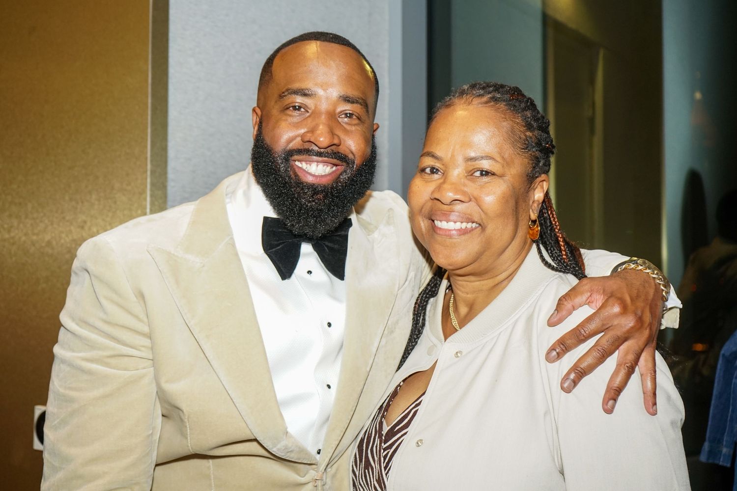 Two people smiling and posing together at a formal event, man in cream tuxedo and woman in black outfit.