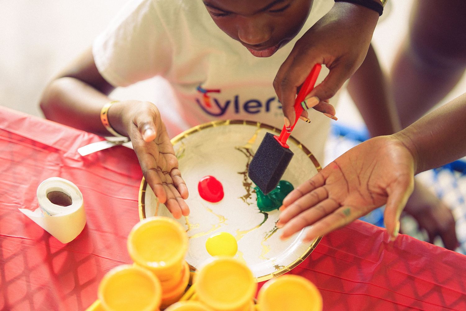 Child in Tyler shirt painting colorful artwork on a paper plate with a brush, surrounded by paint cups on a red table.