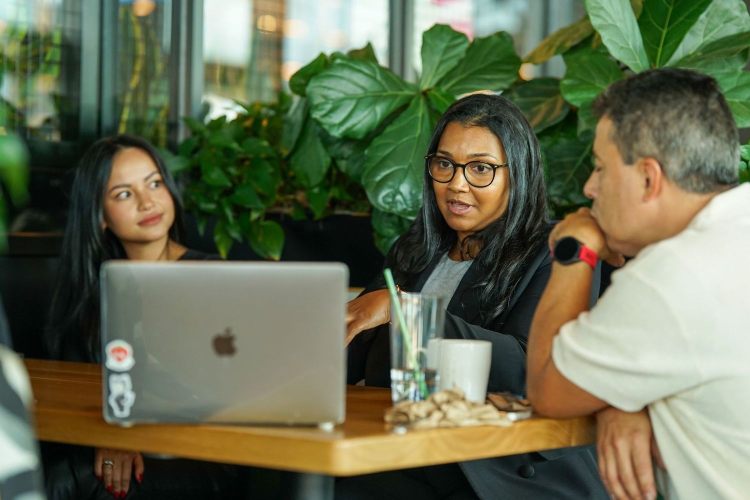Two women and a man having a business meeting at a wooden table with a MacBook in a modern cafe.
