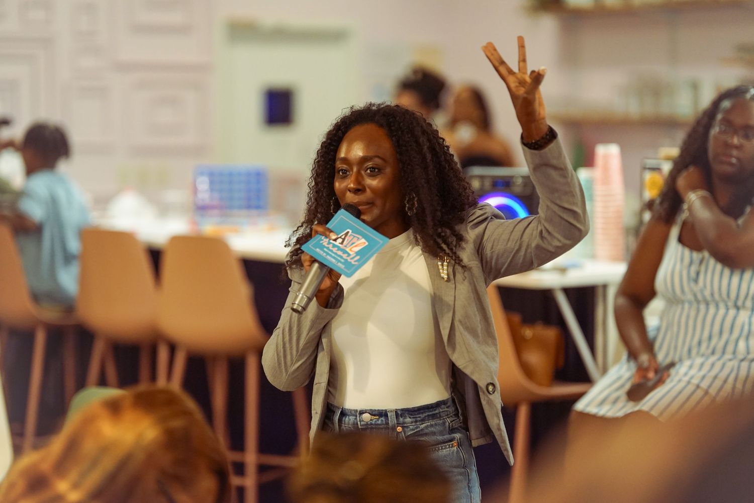 Young Black woman speaking at event, holding microphone and making peace sign, wearing gray blazer and jeans.
