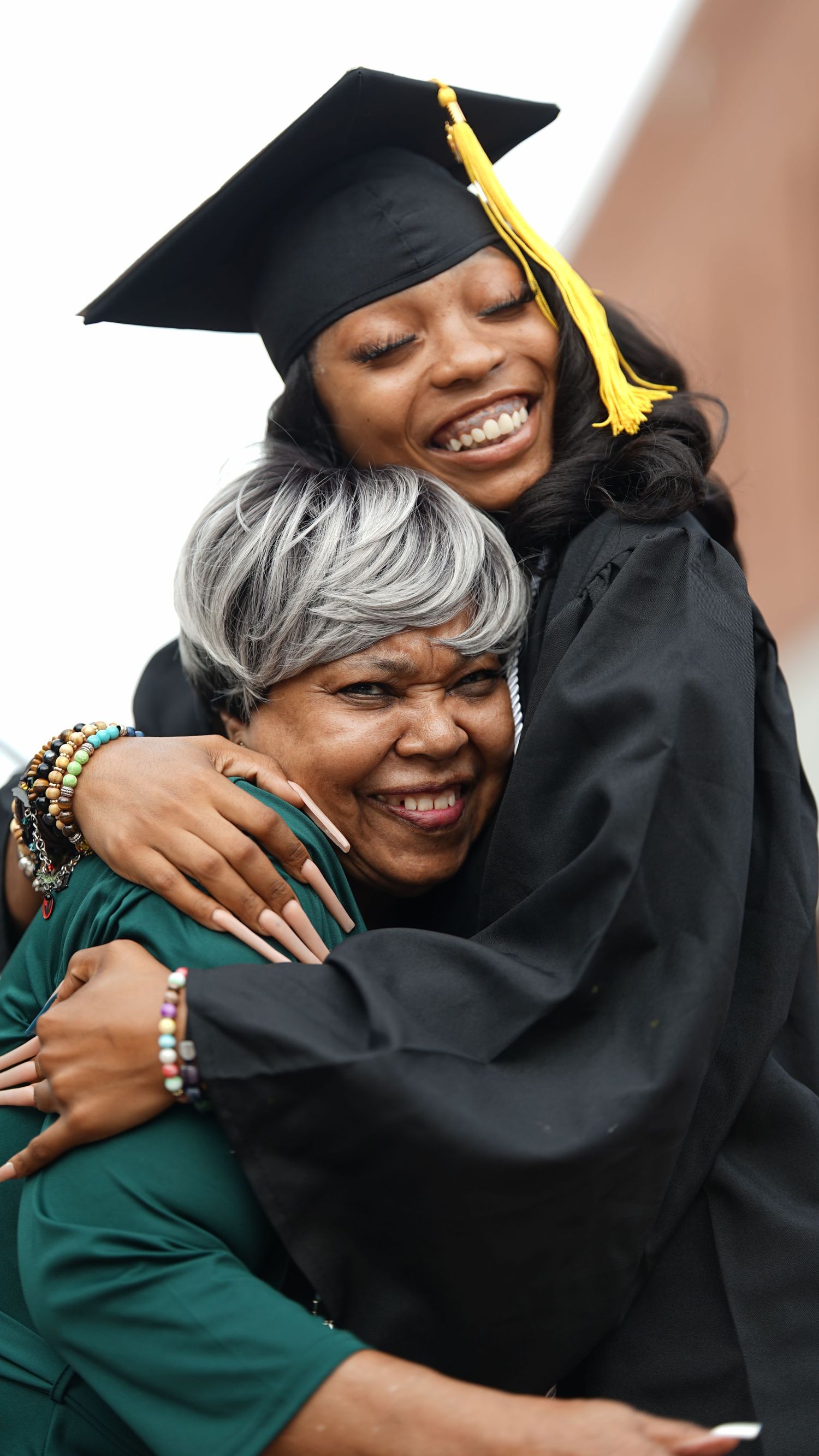 Emotional embrace between graduate in cap and gown and supporter in green.