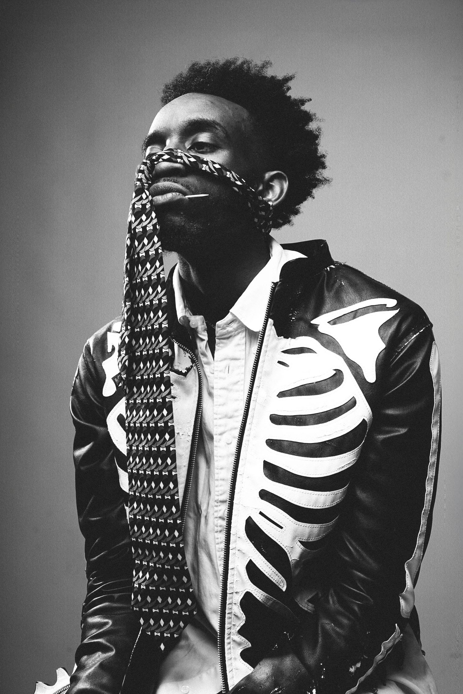 Black and white portrait of a man in skeleton jacket holding a decorative tie over his face with eyes closed.