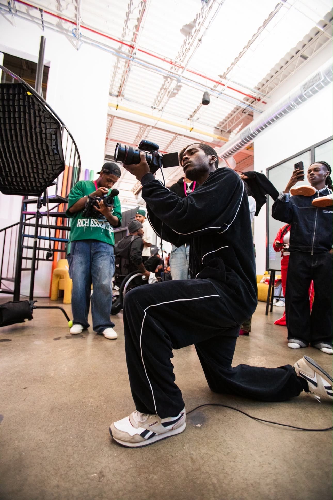 Photographer kneeling with camera at indoor event, surrounded by other photographers and attendees in a warehouse space.
