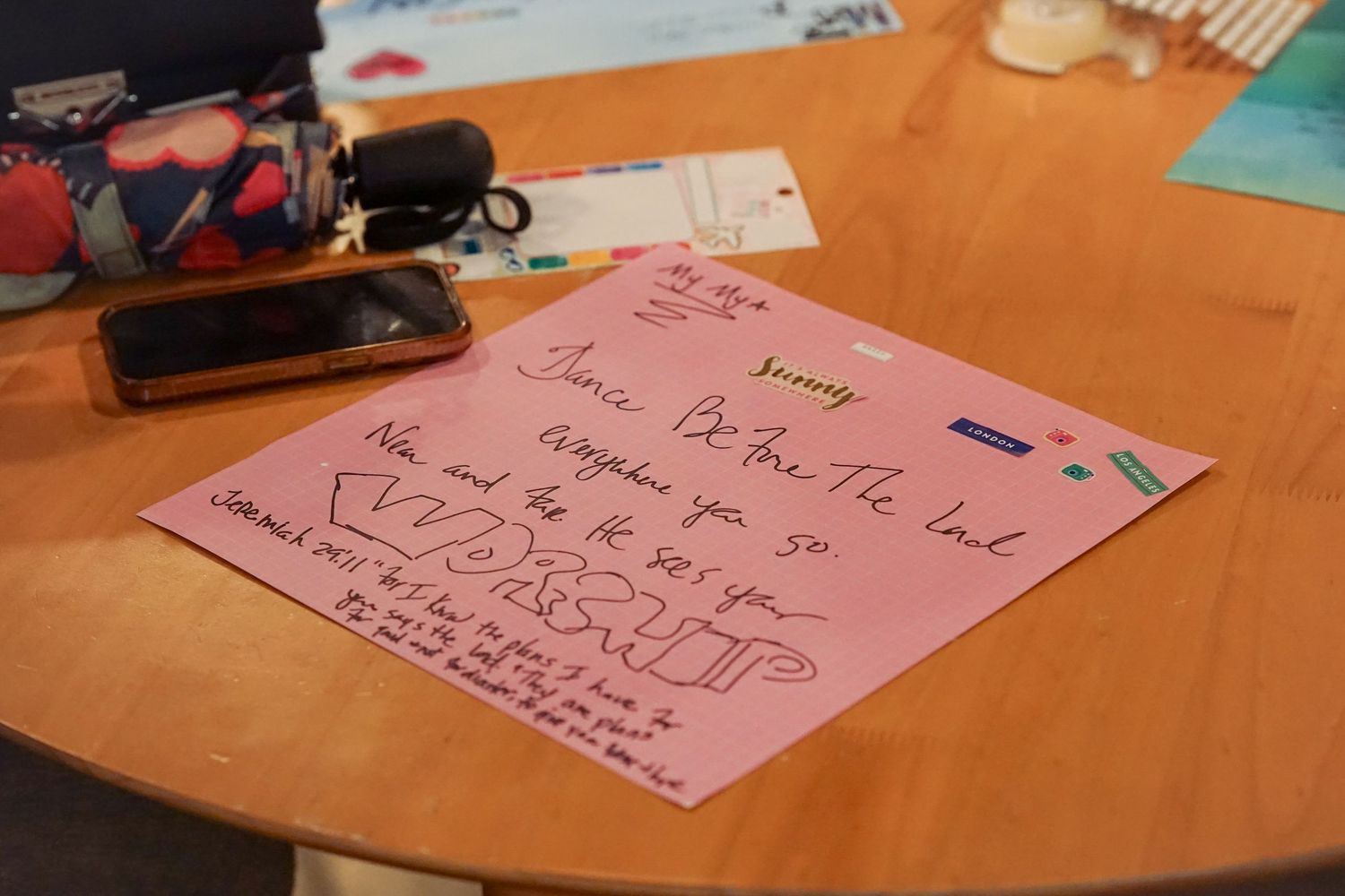 Pink handwritten note on a wooden table with a phone and colorful items in the background.