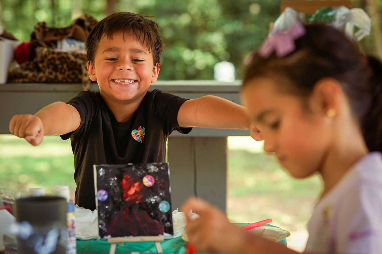 Smiling boy reaches across table during arts and crafts activity outdoors at Tyler's Way Foundation event.