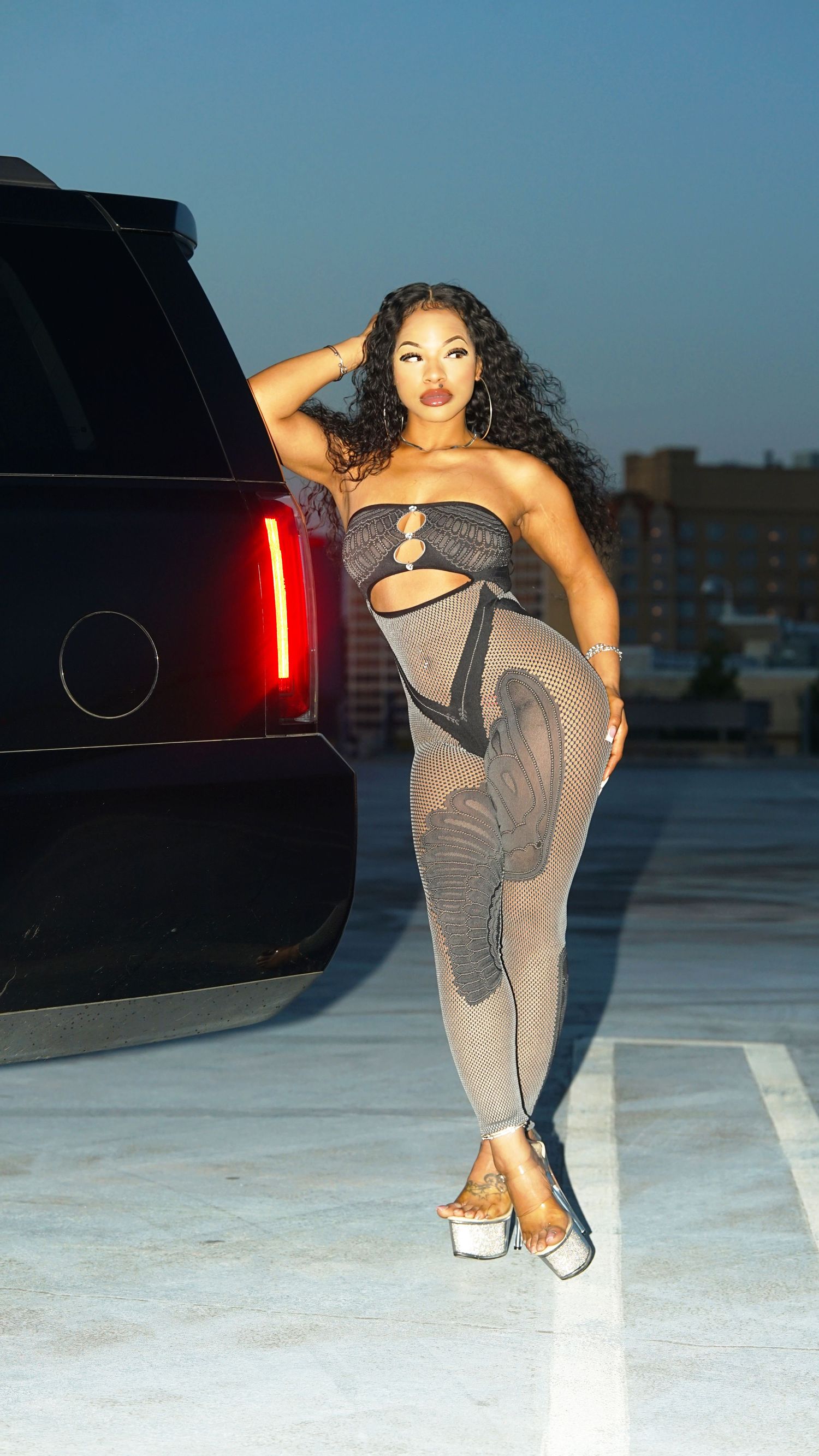 Fashion model in grey evening wear poses dramatically by black SUV at dusk.