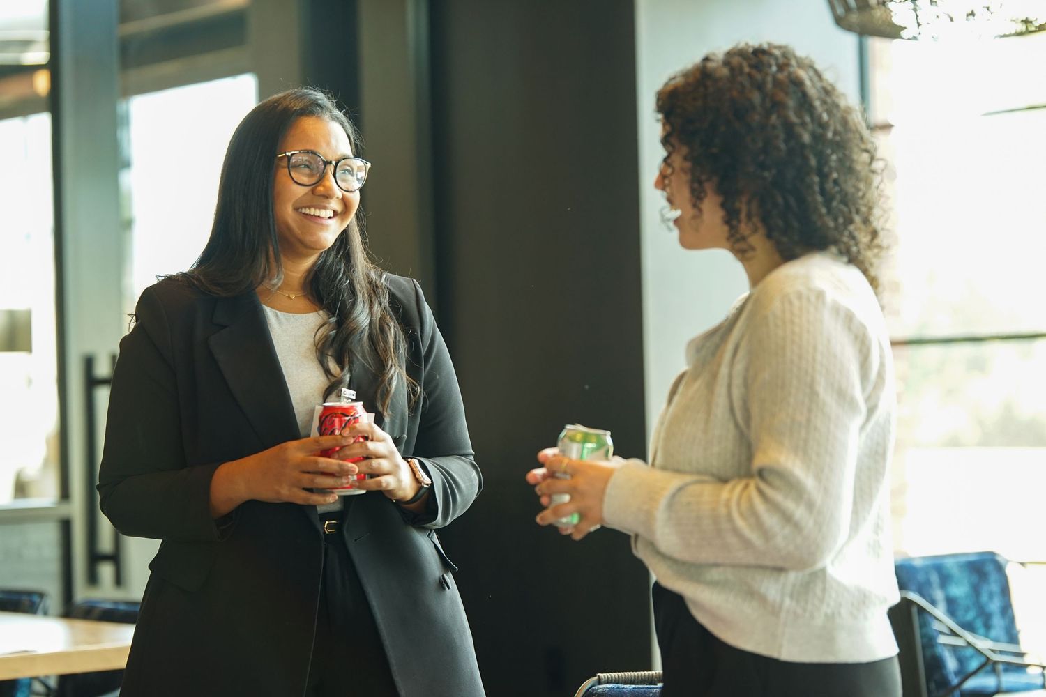 Two businesswomen smile and chat while holding drinks in a bright modern office.