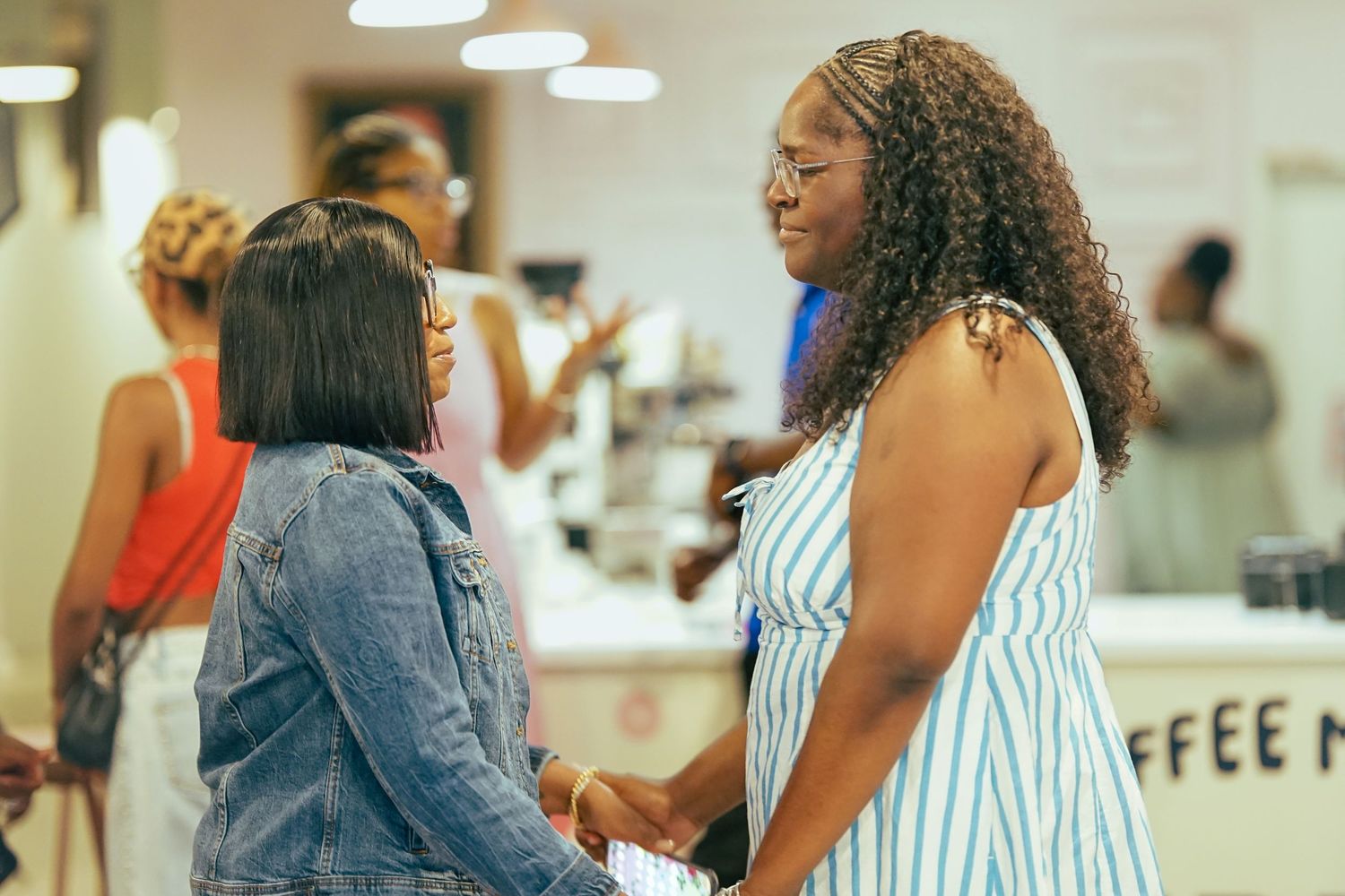 Two Black women smiling and holding hands in conversation inside a coffee shop.