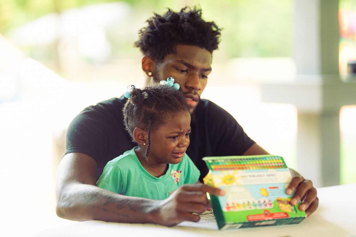 Young man reads educational book with toddler girl on his lap at outdoor community event.