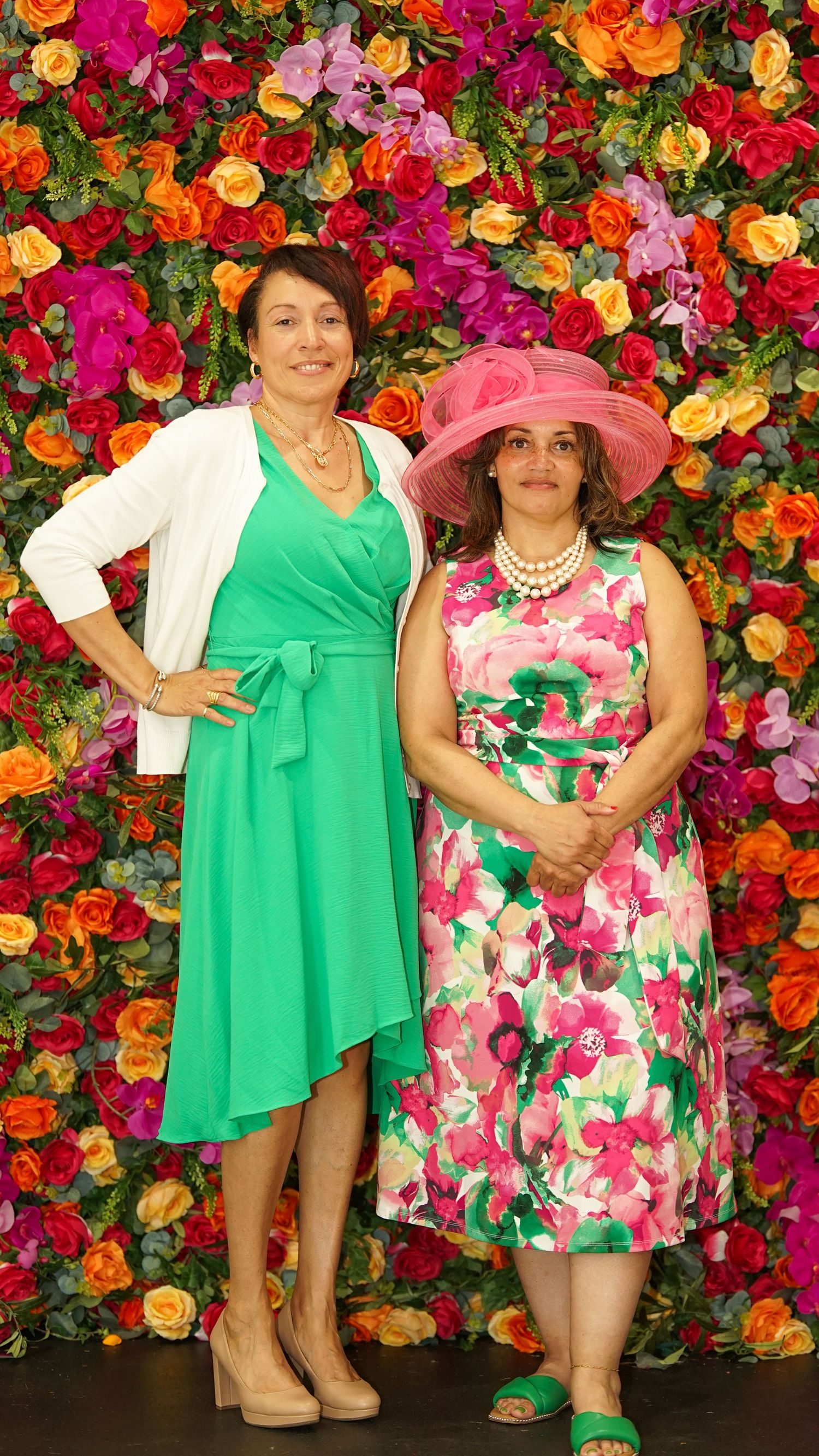 Two people pose in colorful dresses against a vibrant floral wall backdrop at an elegant event.