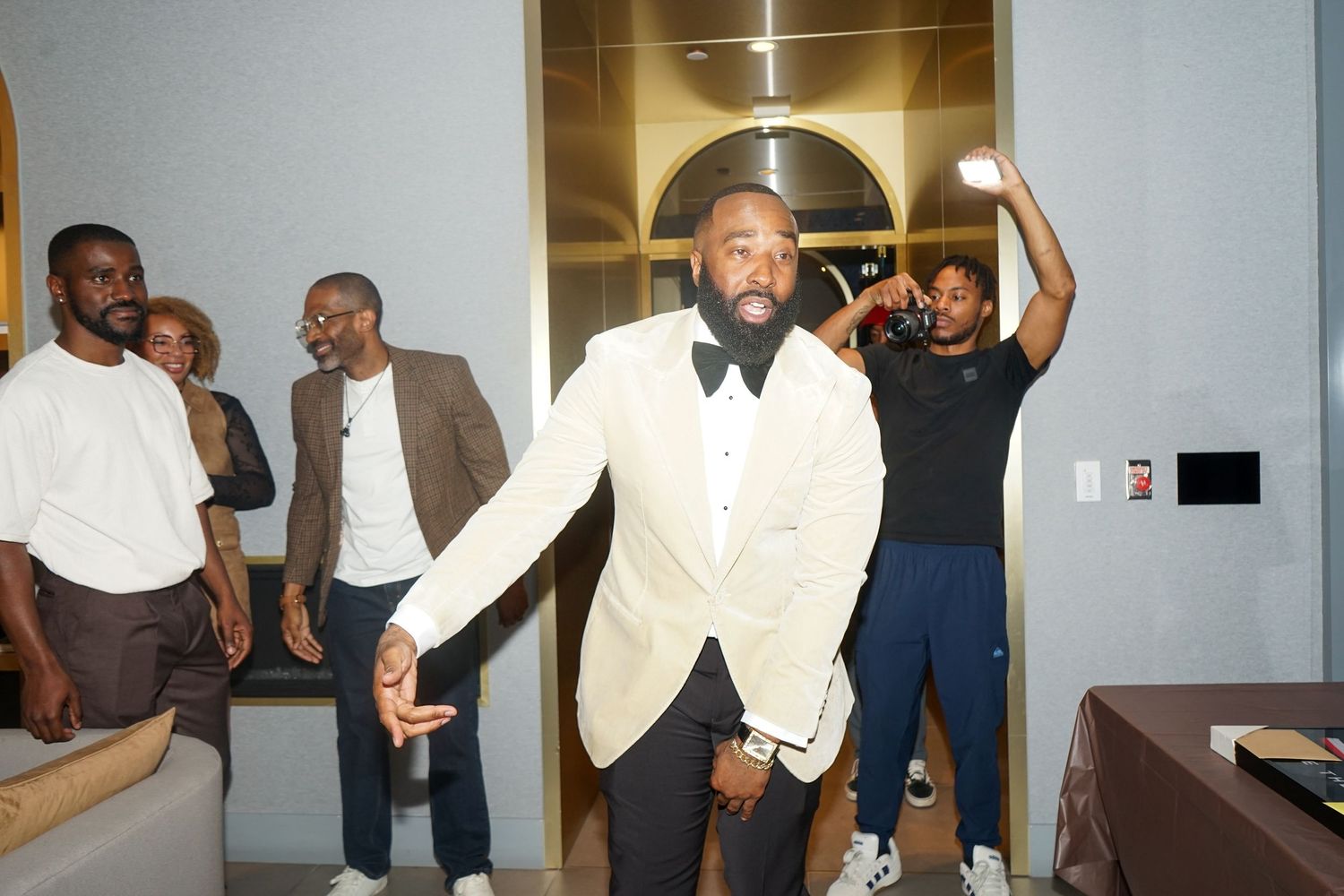 Man in white tuxedo jacket dancing at a party with friends gathered in a modern room.