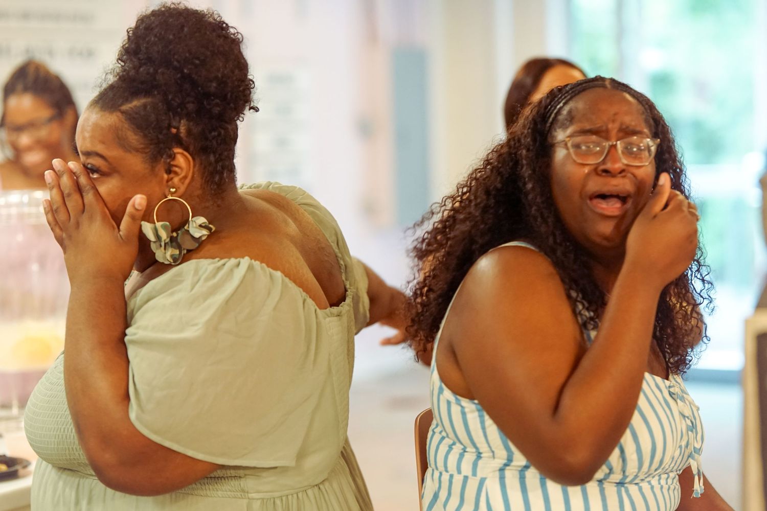 Two Black women cry and cover their faces with their hands at an emotional gathering.