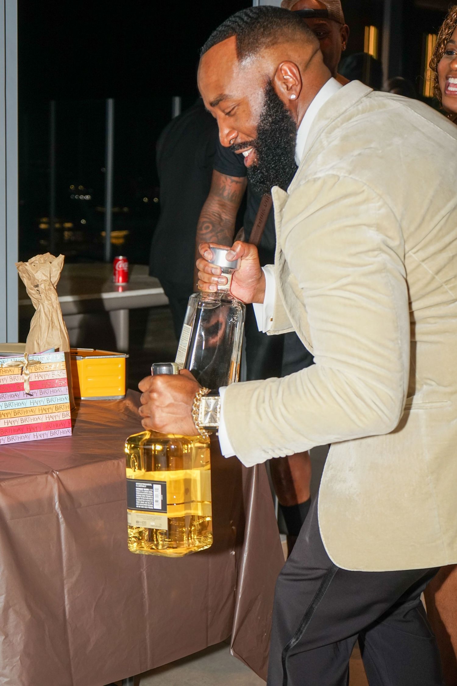Man in white velvet blazer pouring large bottle of liquor at an elegant evening event.