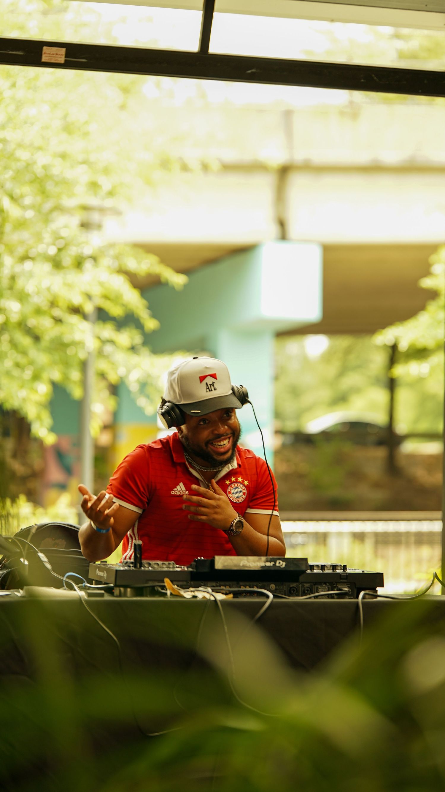 DJ in red jersey and cap operates mixing equipment at an outdoor event.