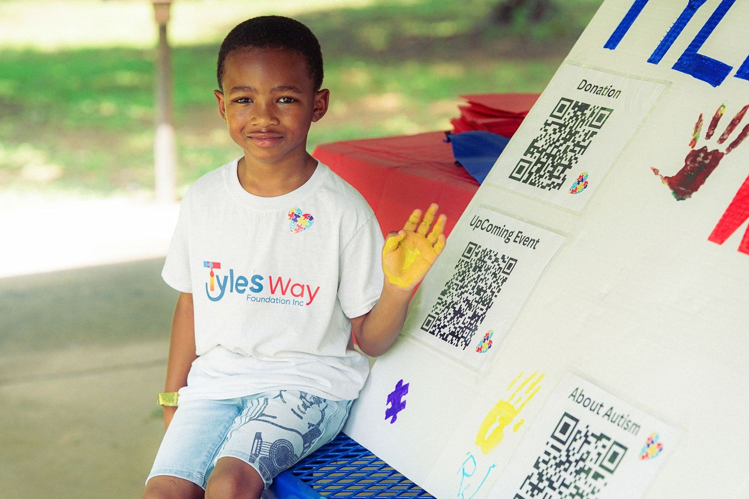 A smiling boy in a Kyle's Way t-shirt sits beside a colorful event display board with QR codes.