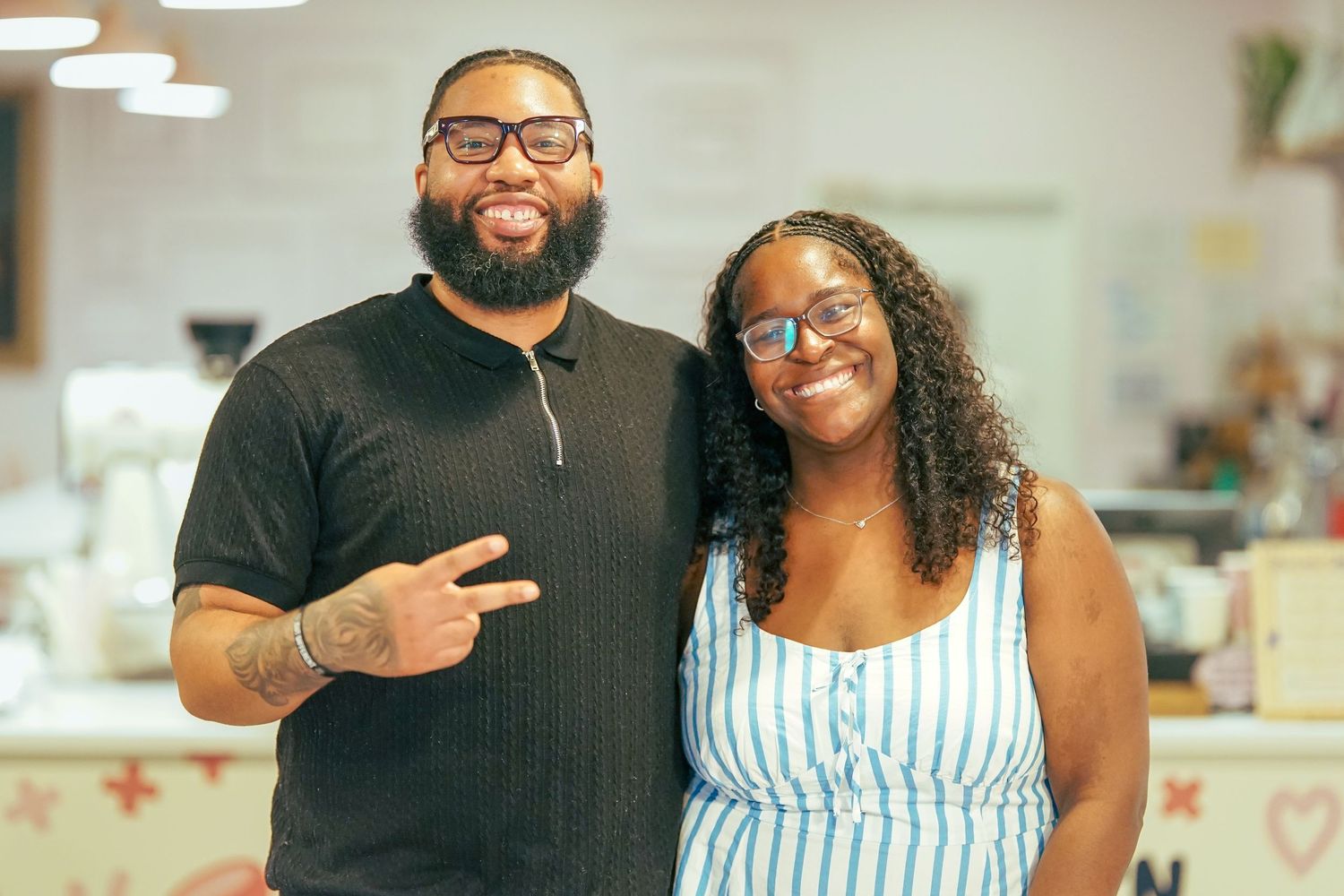A bearded Black man and a smiling woman in glasses pose together making a peace sign.