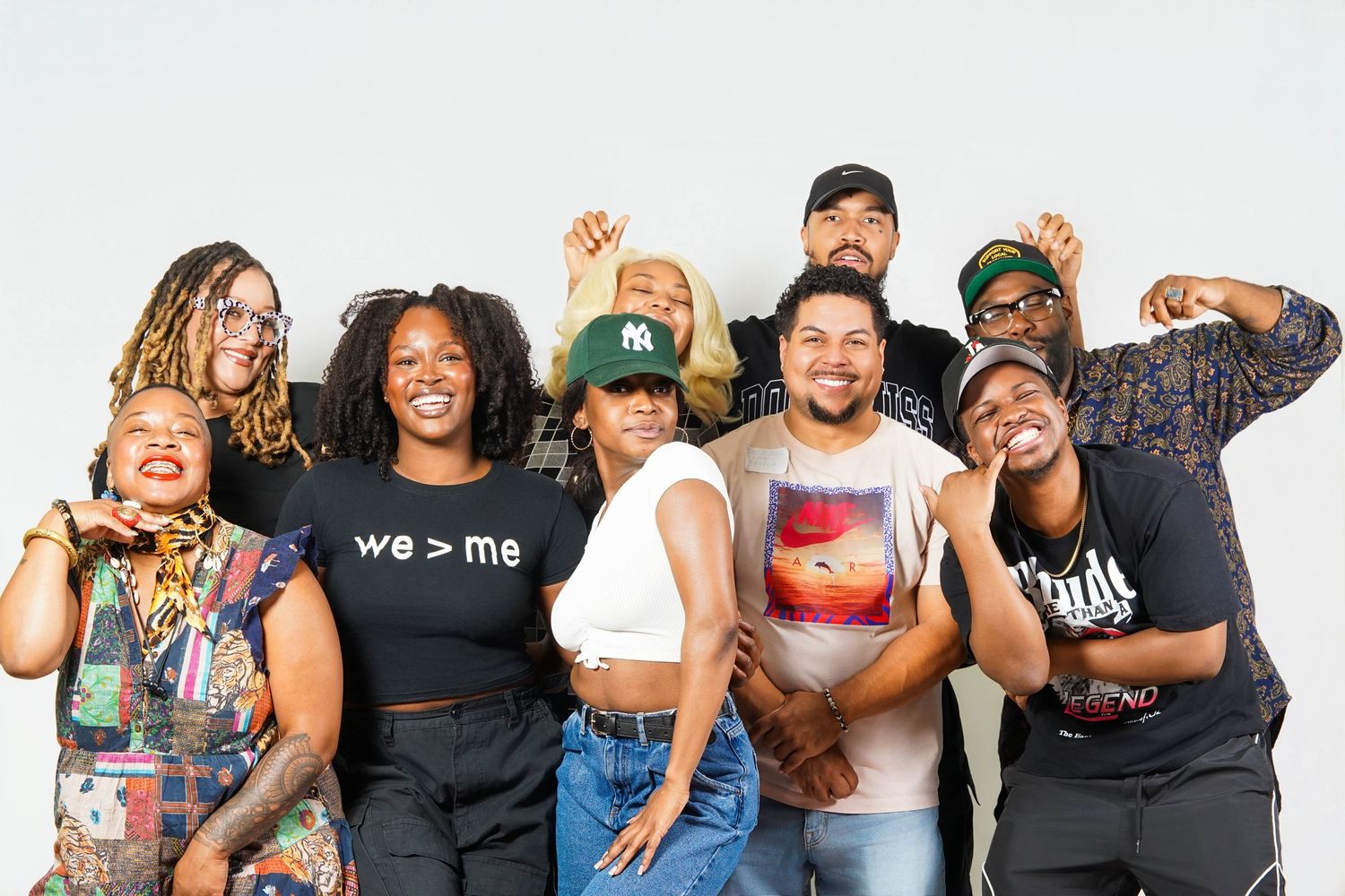 Diverse group of eight friends posing together, smiling and laughing, one wearing a 'we > me' shirt.