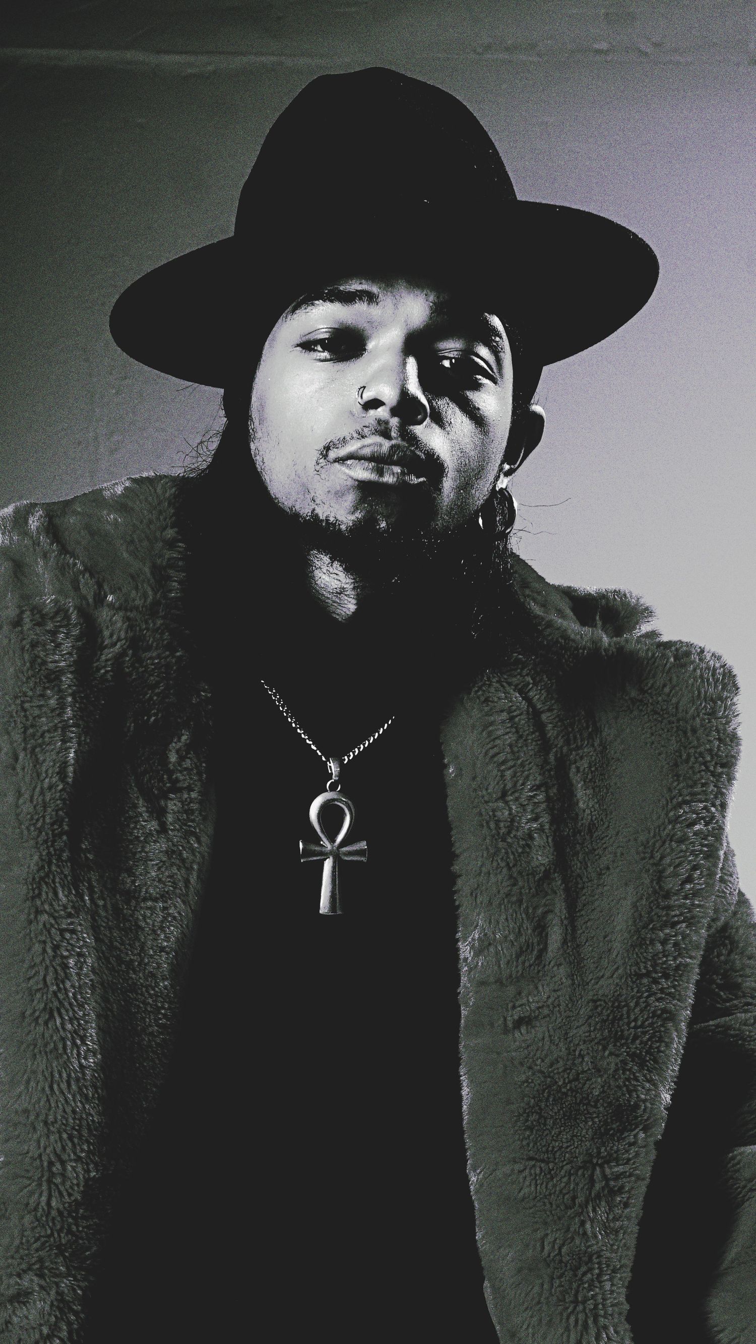 Dramatic black and white portrait of person wearing hat and ankh necklace.