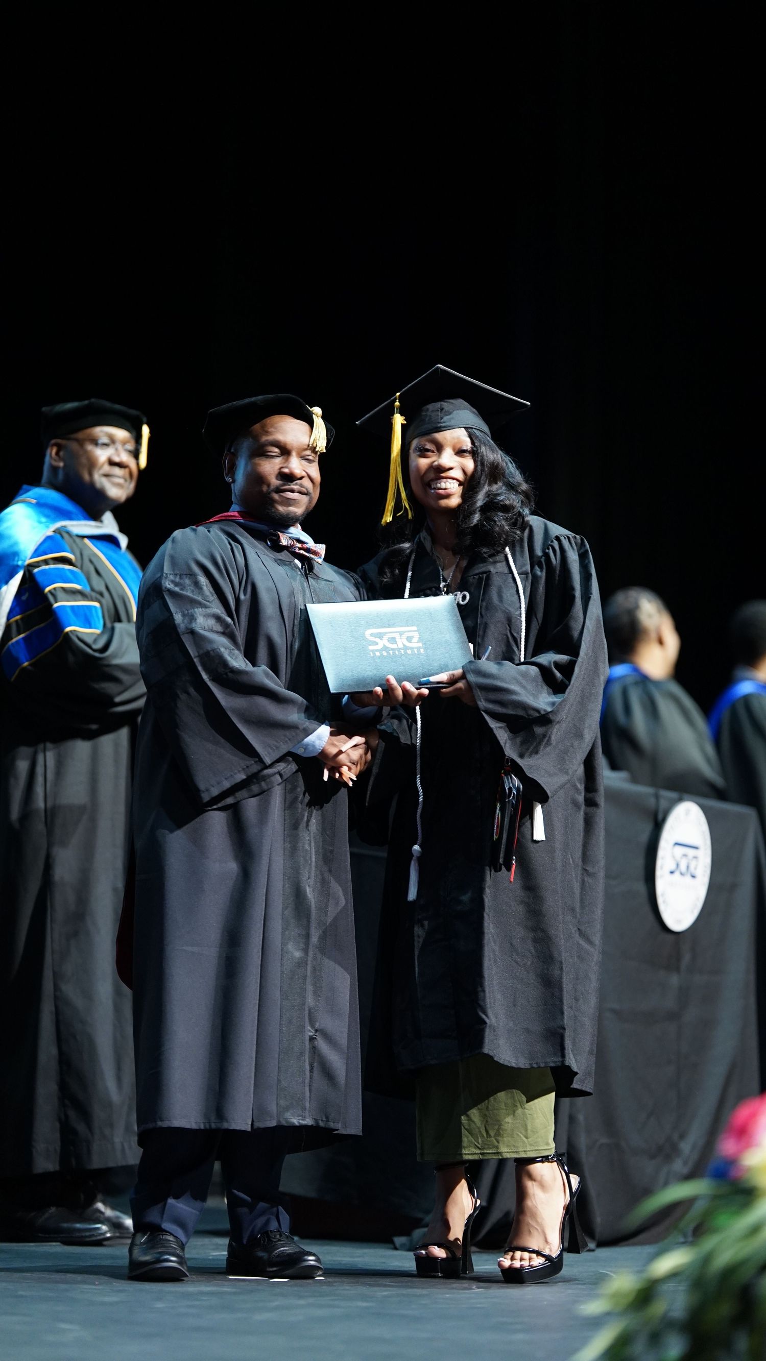 Graduates in black academic regalia receive diplomas during a formal graduation ceremony.
