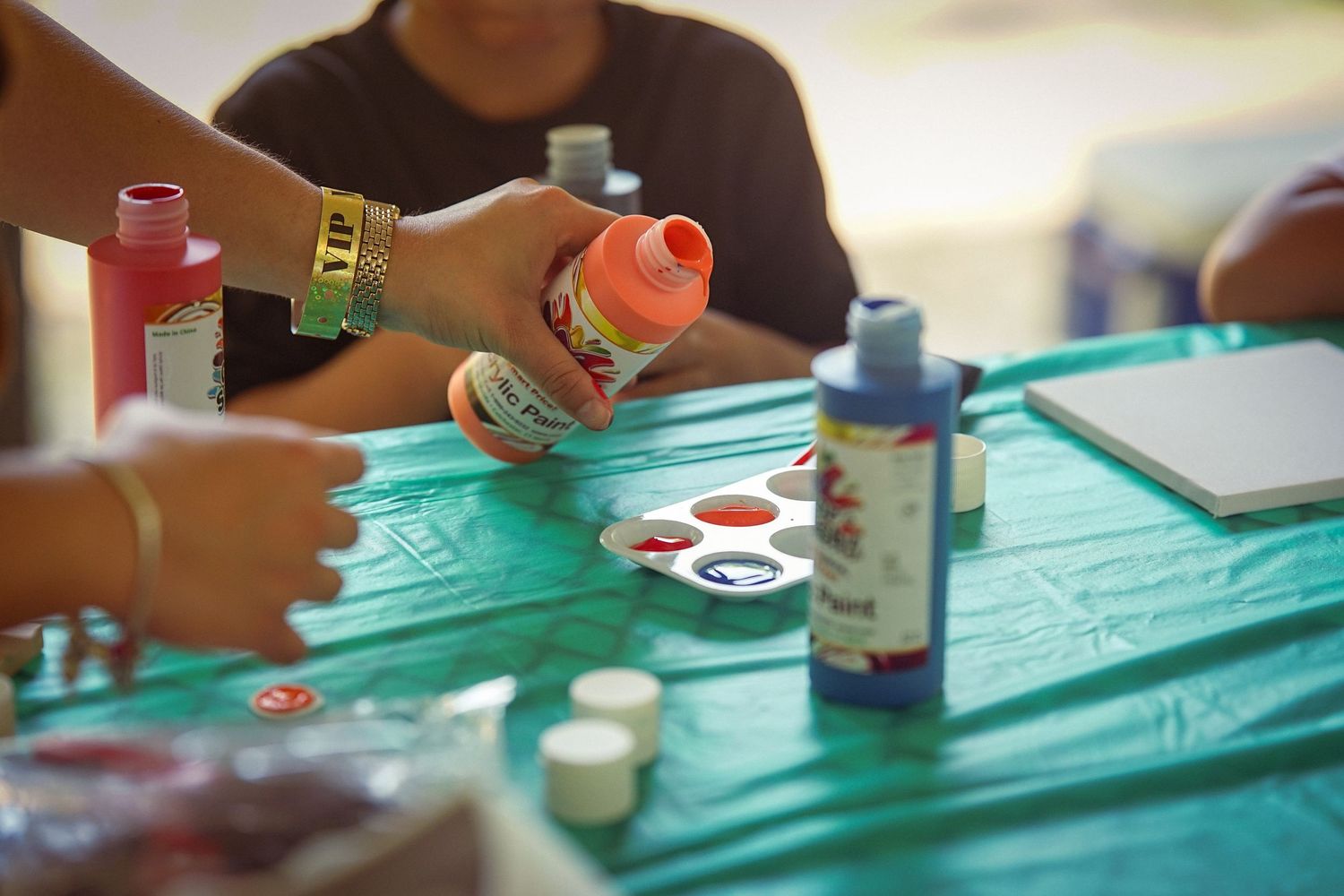 Children doing arts and crafts with colorful paint bottles and a palette on a teal table.