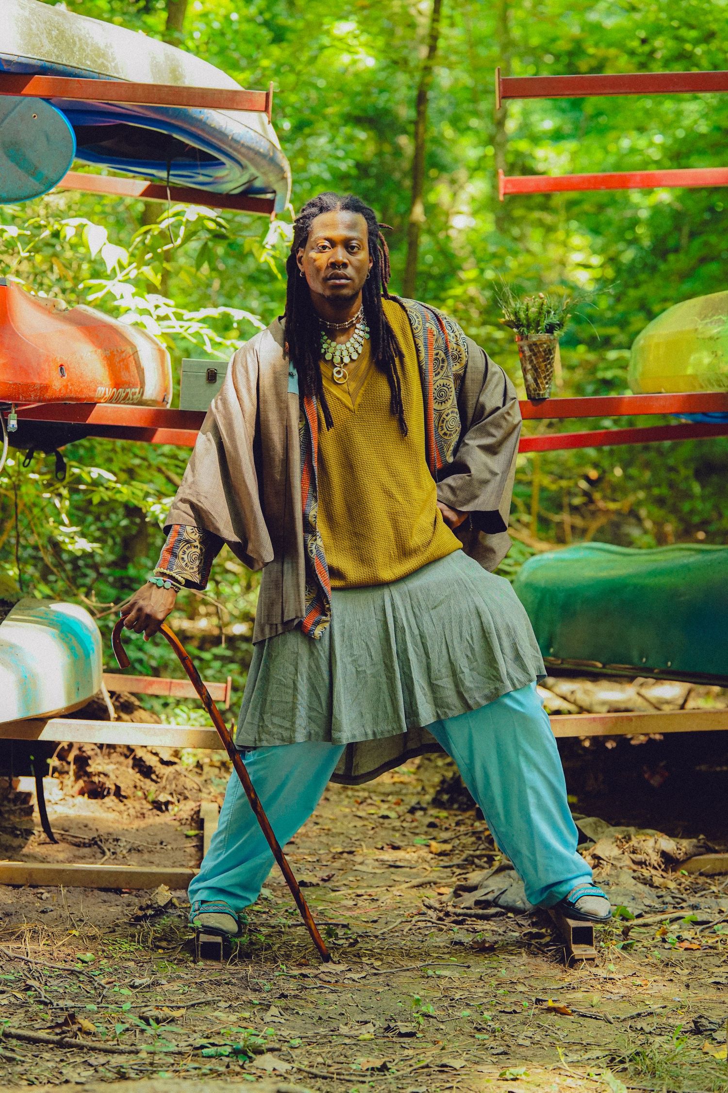 Man with dreadlocks in eclectic layered outfit holding a wooden cane, posing near colorful kayaks in a forest.