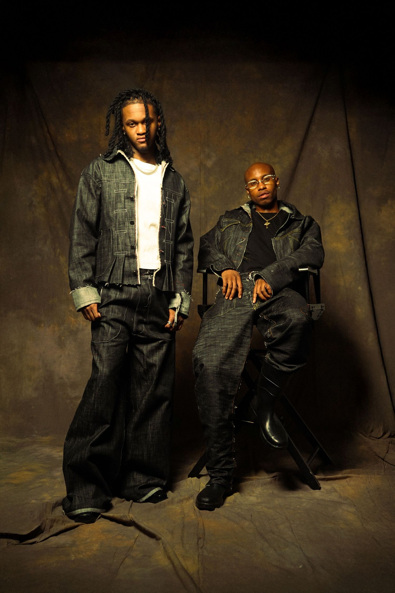 Two performers pose against a dark backdrop wearing denim and casual urban streetwear in a studio portrait setting.