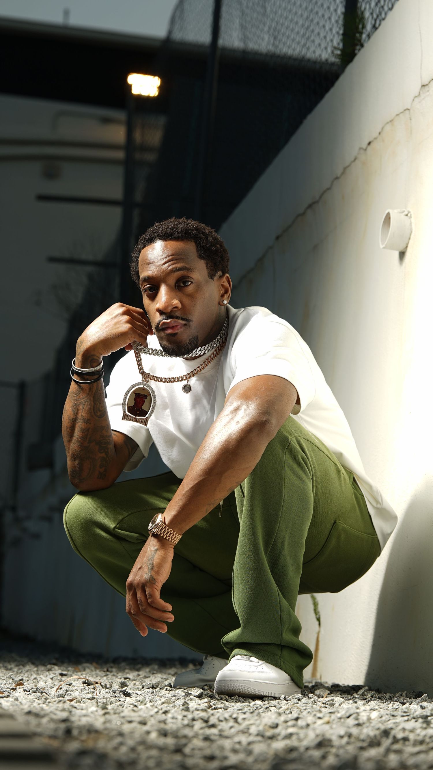Athletic man in white t-shirt and olive pants crouches against wall in urban setting with dramatic lighting.