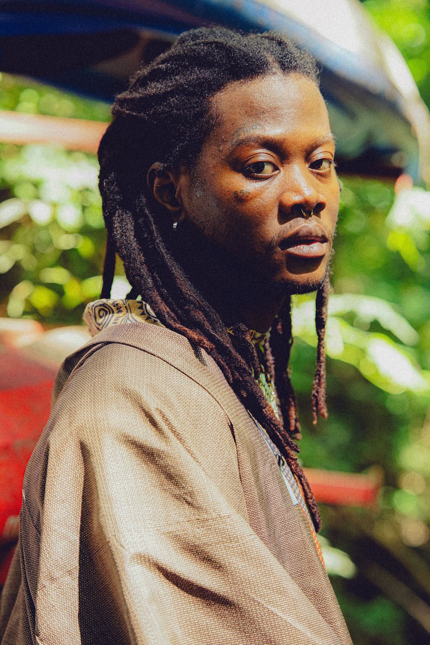 Young Black man with dreadlocks wearing a brown jacket, posing outdoors surrounded by lush green foliage.