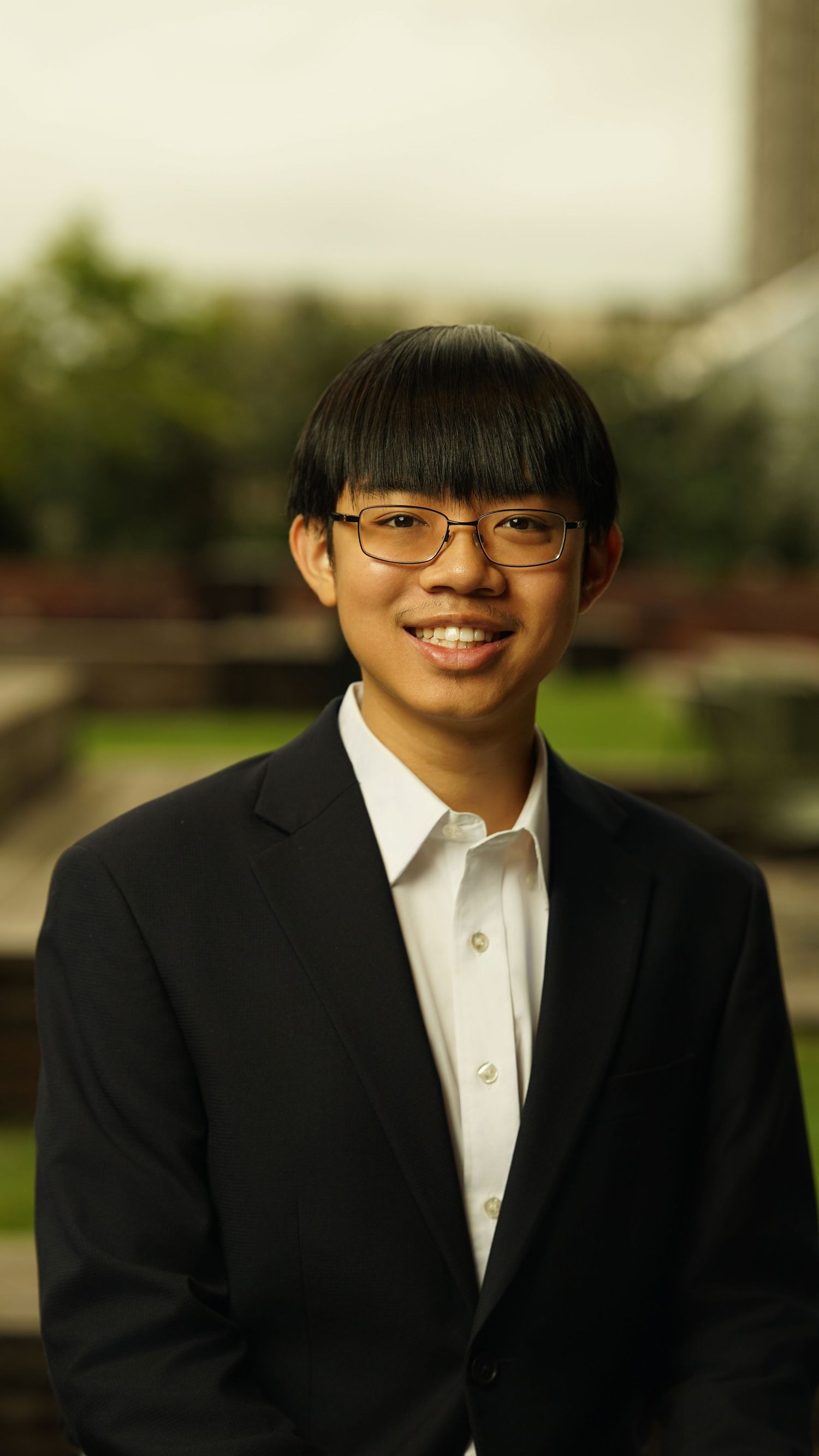 Professional headshot of person in black suit and white shirt wearing glasses with bangs.