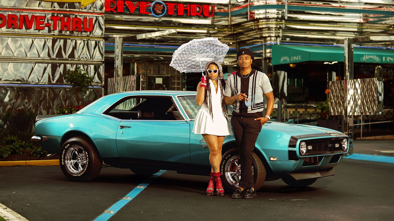 A vintage teal Chevrolet Camaro with two people posing beside it in retro outfits at a drive-in restaurant.