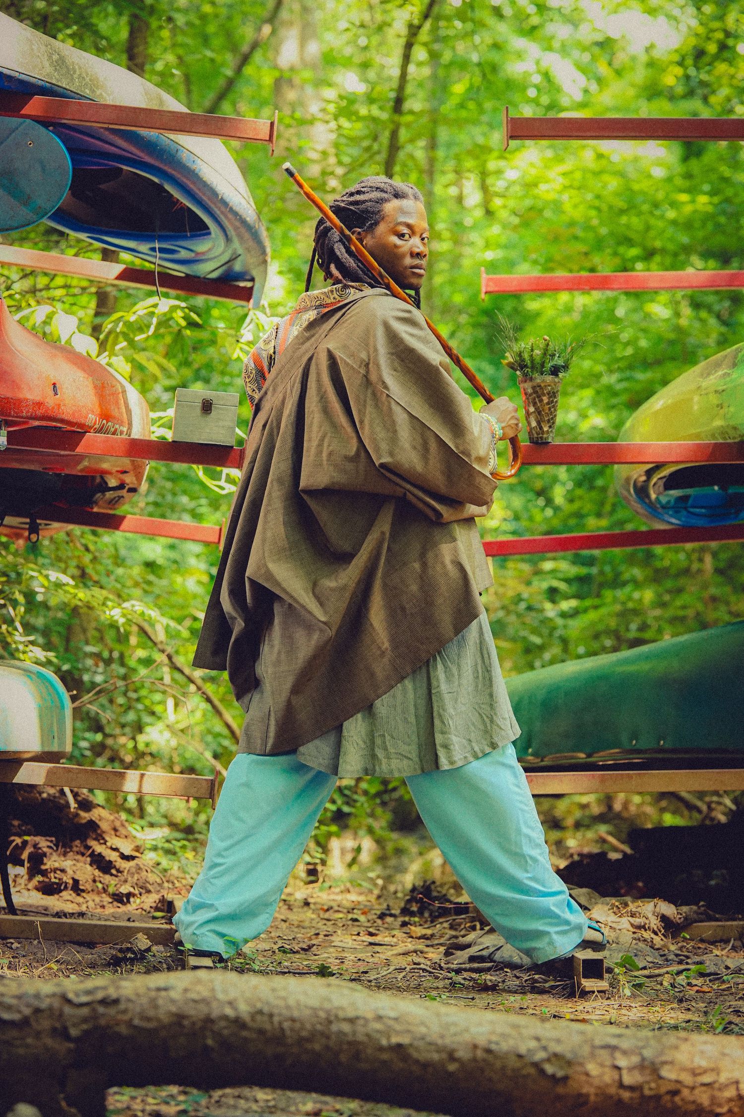 Person with dreadlocks in brown cloak and teal pants stands near colorful kayaks on outdoor storage rack in forest.