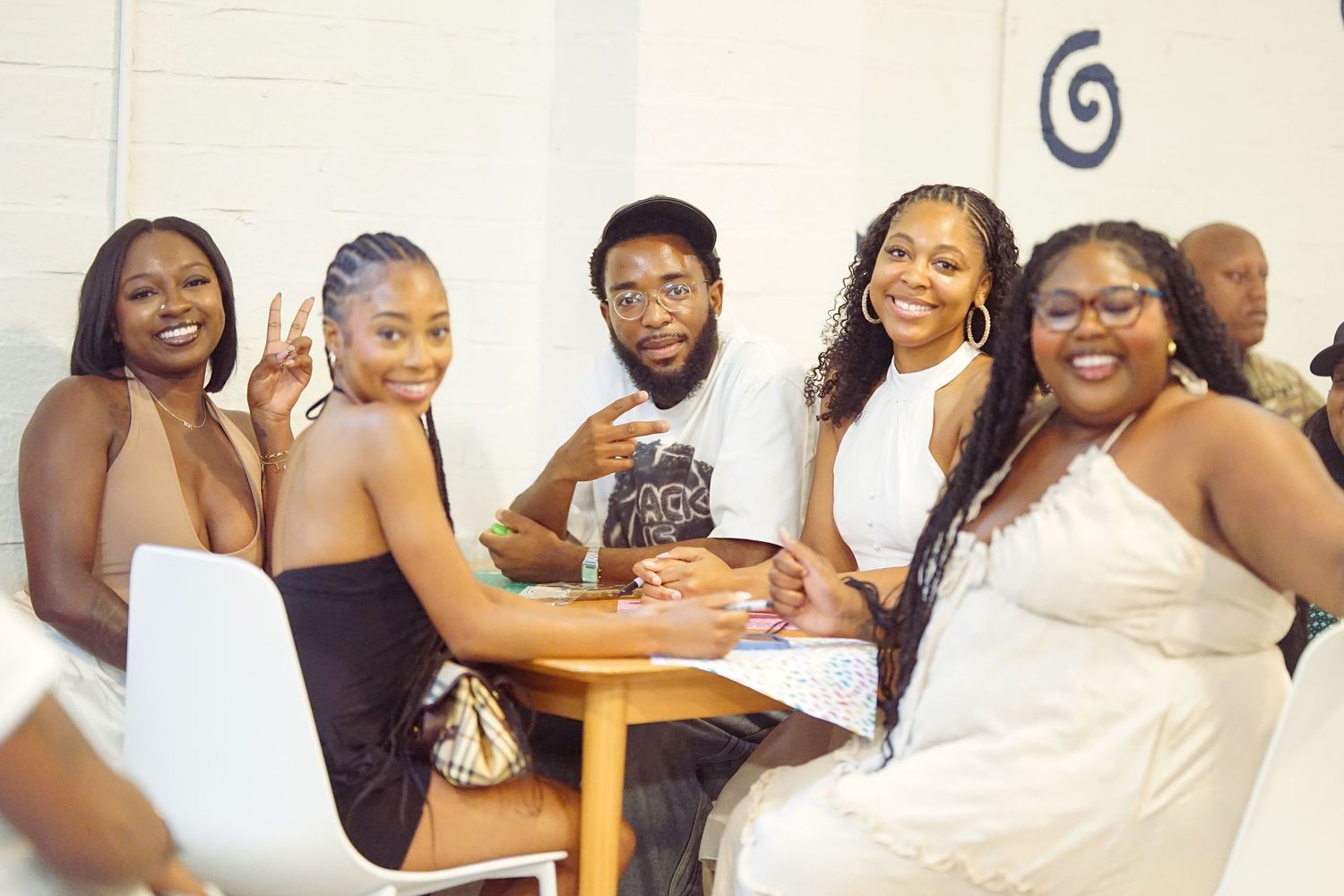 Five friends smiling and posing together at a table in a bright white venue, dressed in stylish outfits.