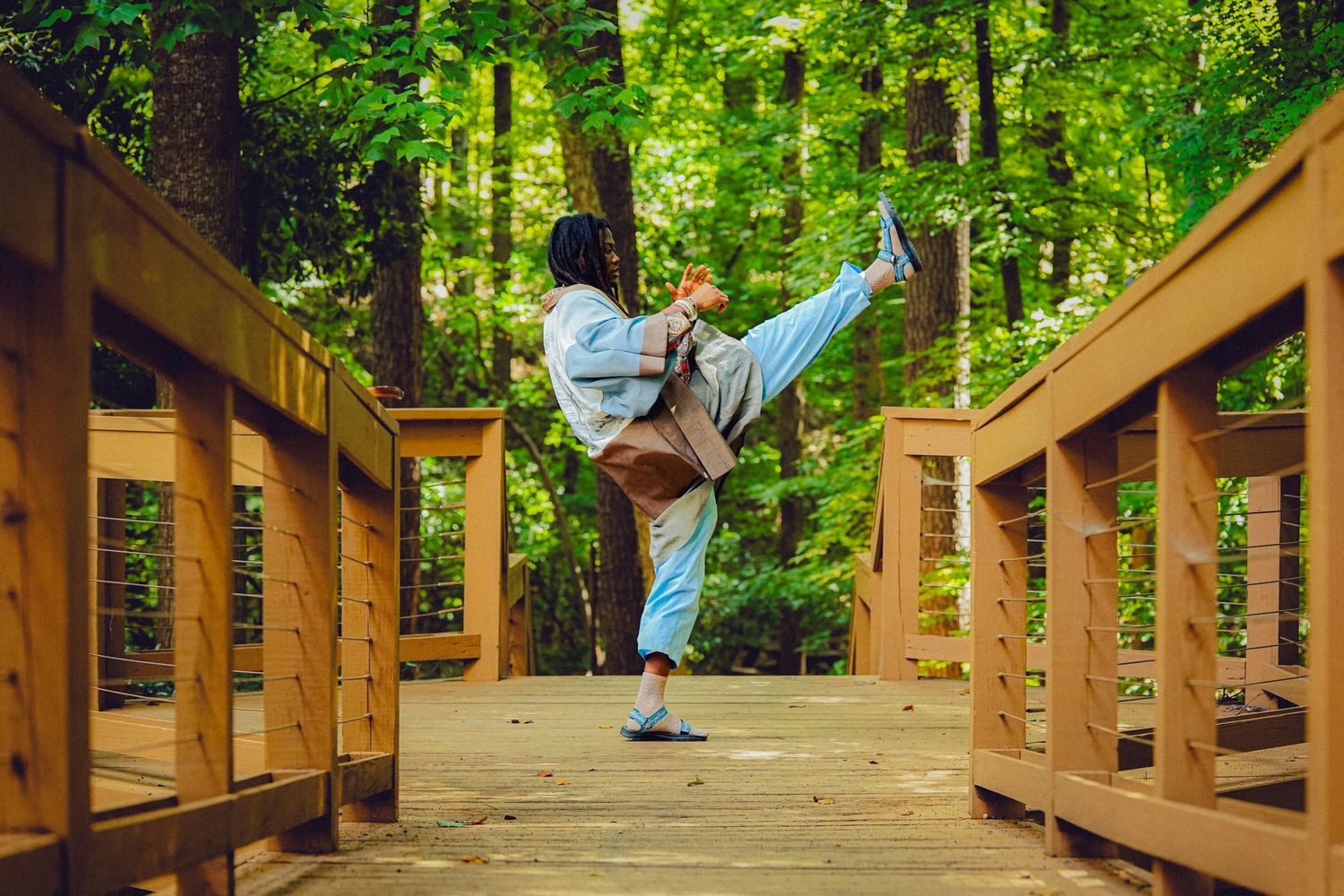 Person performing a high kick martial arts move on a wooden bridge surrounded by lush green forest trees.