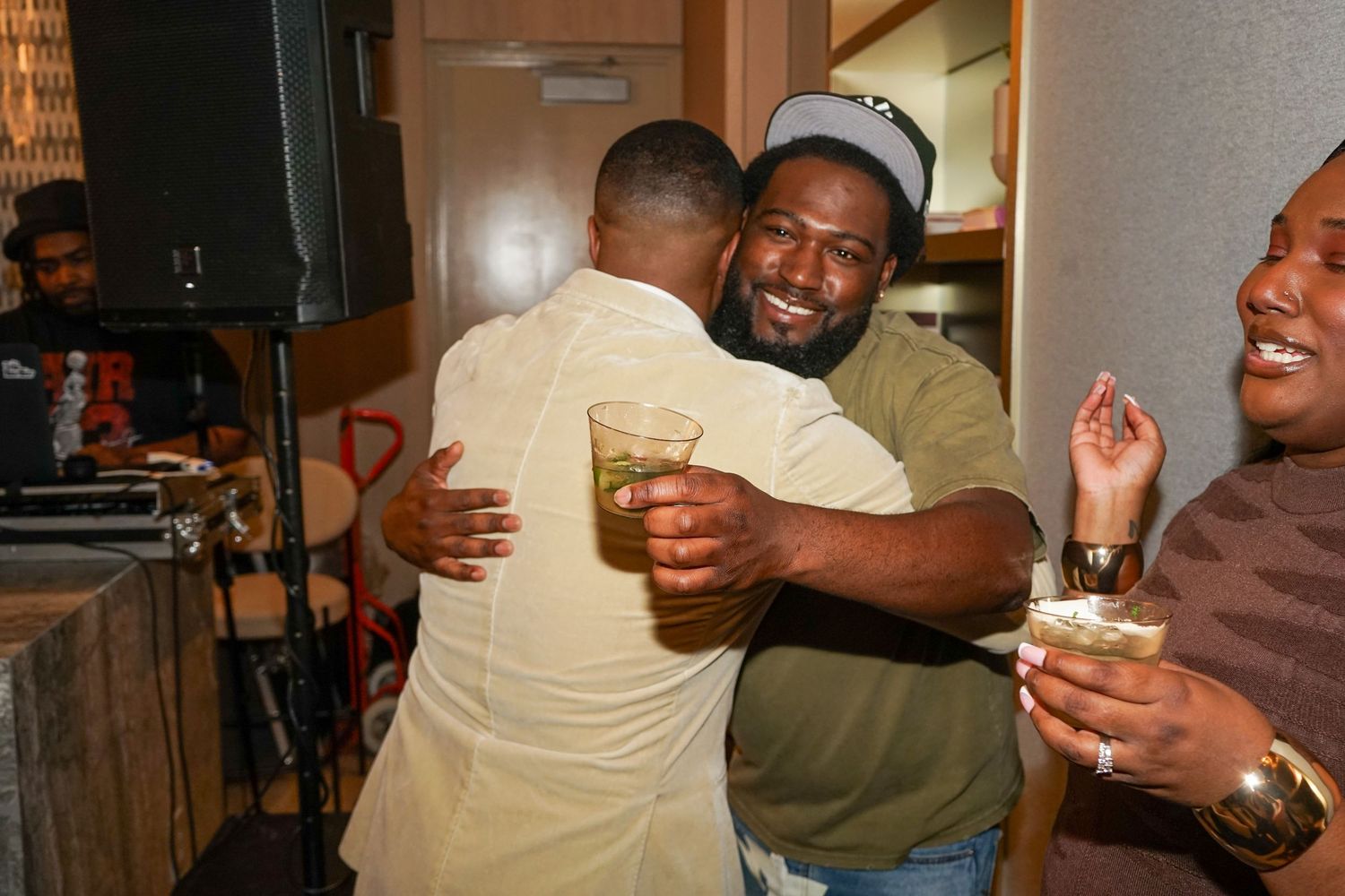 Two men share a warm hug at a lively indoor party, holding drinks while others celebrate around them.