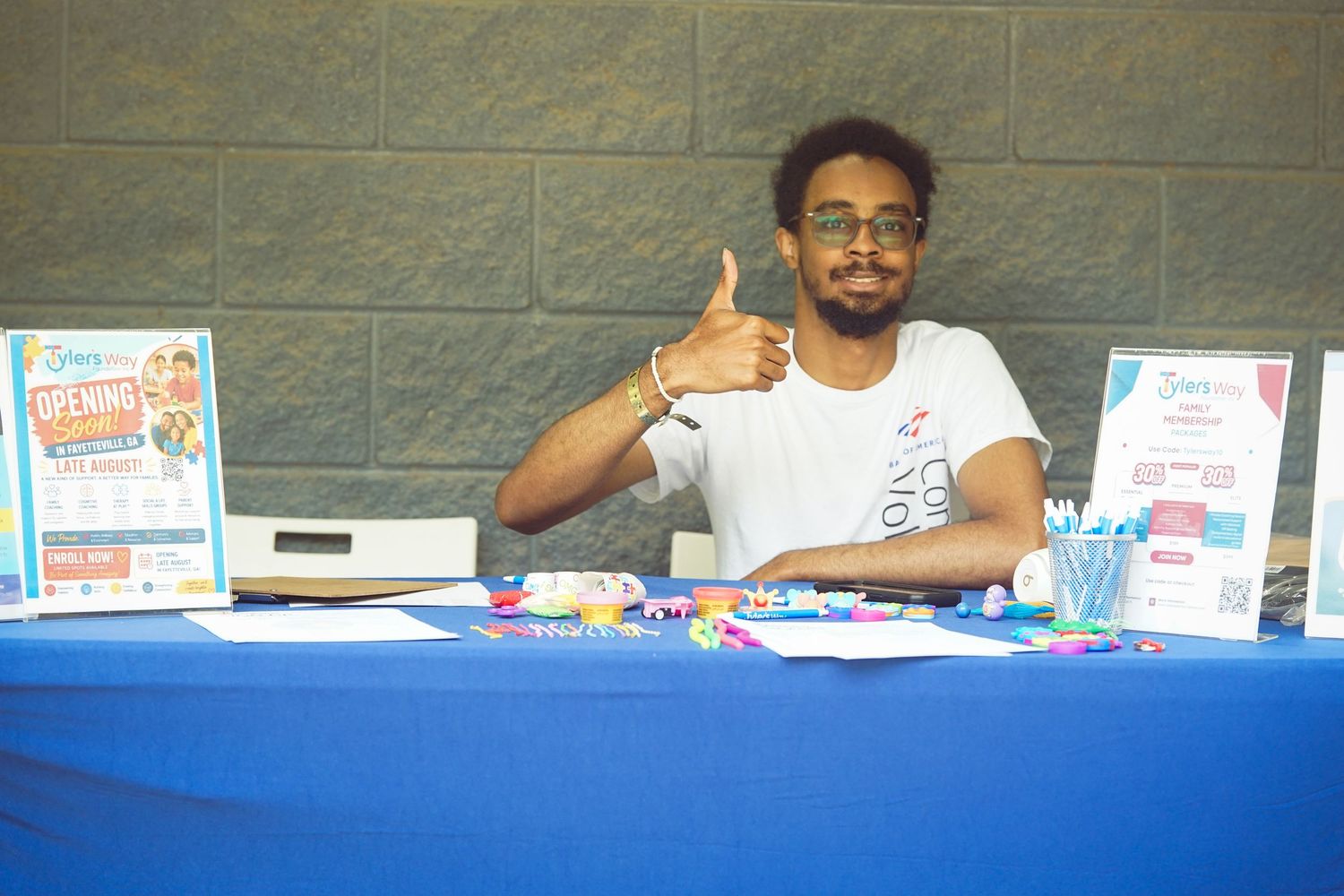 Smiling young man giving thumbs up at a blue table with school supply drive promotional materials.