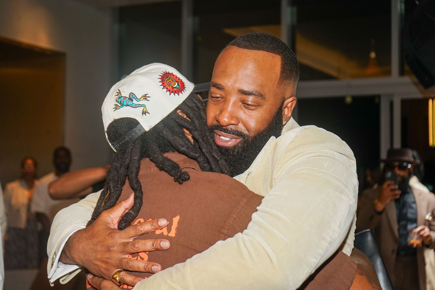 Two men sharing a warm hug at an indoor event, one wearing a decorative cap and dreadlocks.