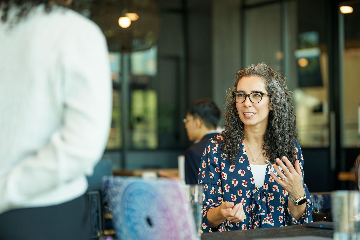 Woman with curly hair and glasses engaged in conversation at an outdoor cafe setting.