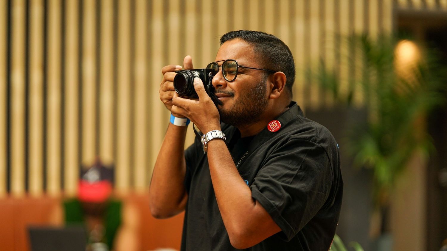 Person wearing glasses photographs an event against a wooden fence background.