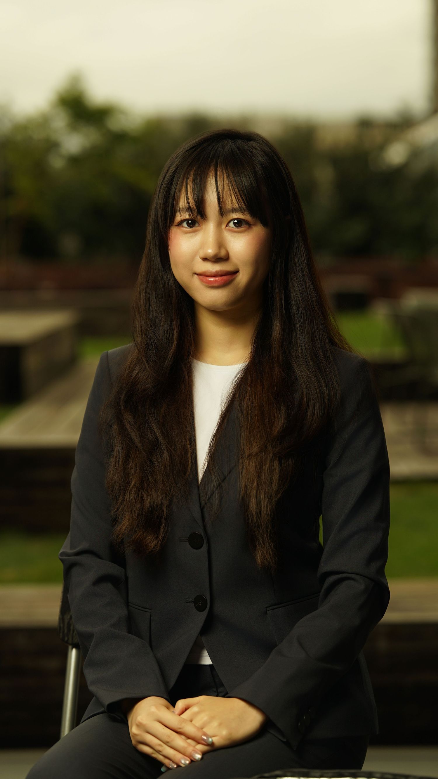Business portrait of person in black blazer and white top with bangs against natural backdrop.