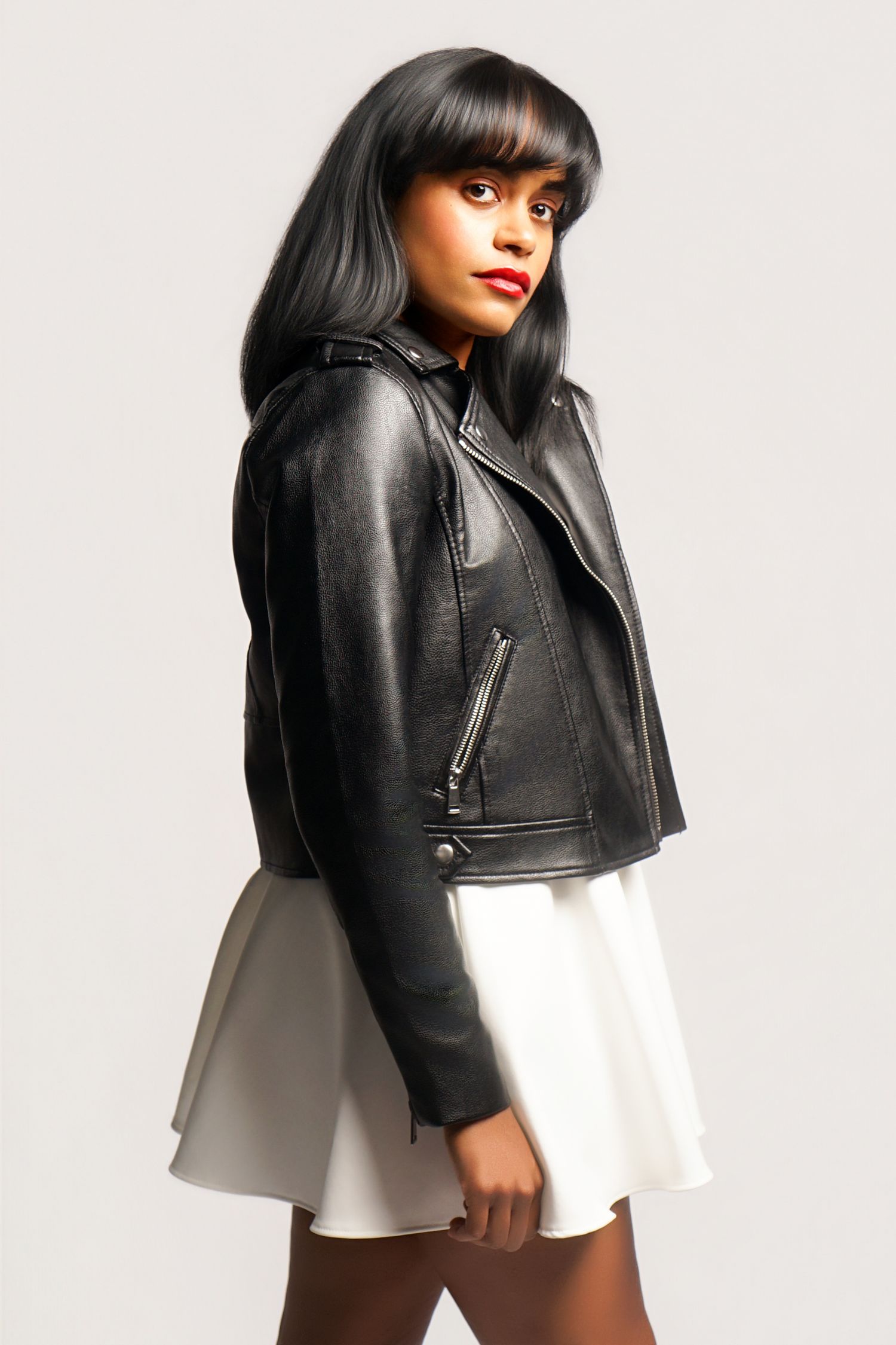 Woman in black leather biker jacket and white mini skirt with red lips and black hair poses confidently.