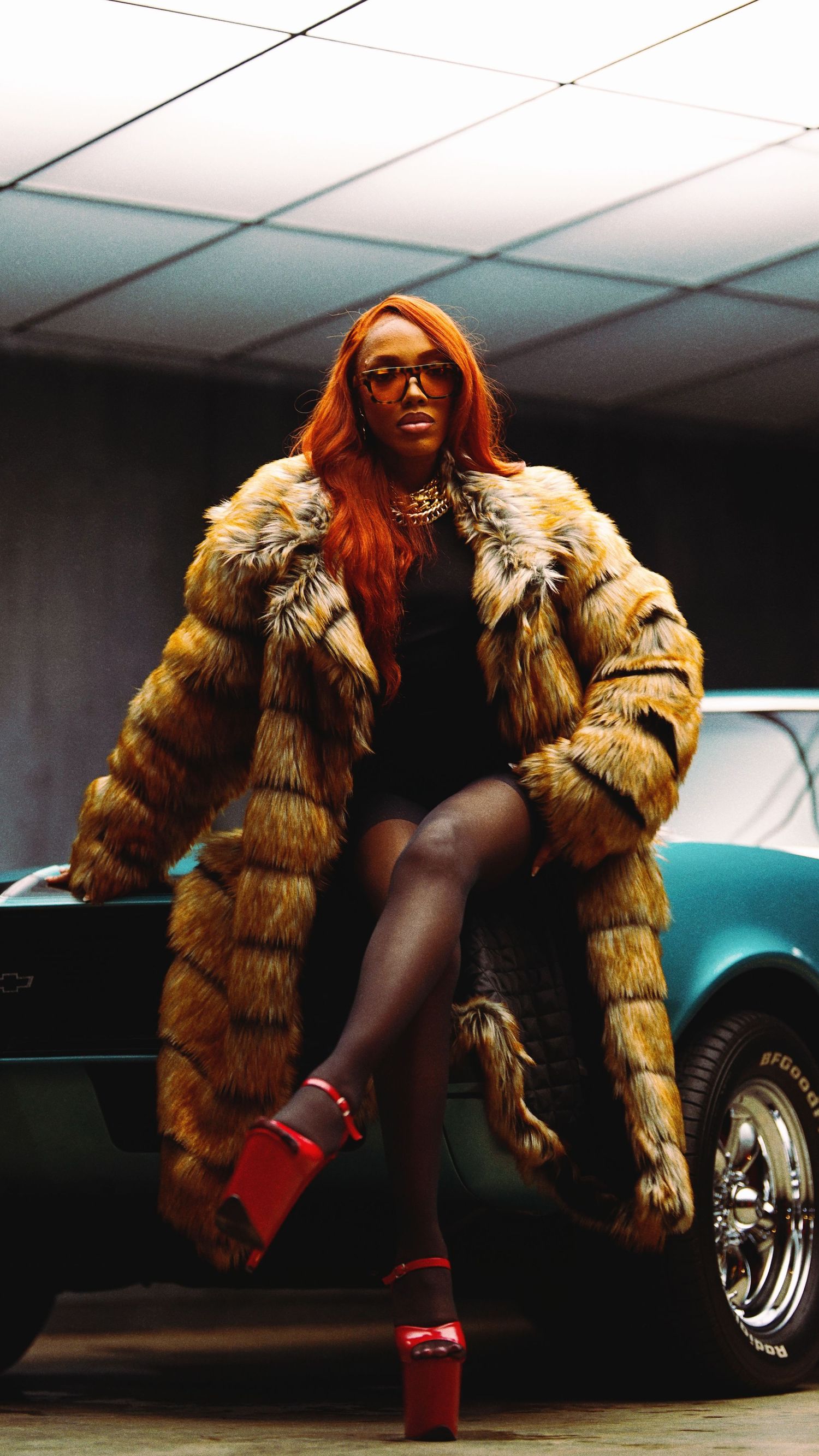 Fashion model in a luxurious golden fur coat and red boots poses in front of a vintage car.