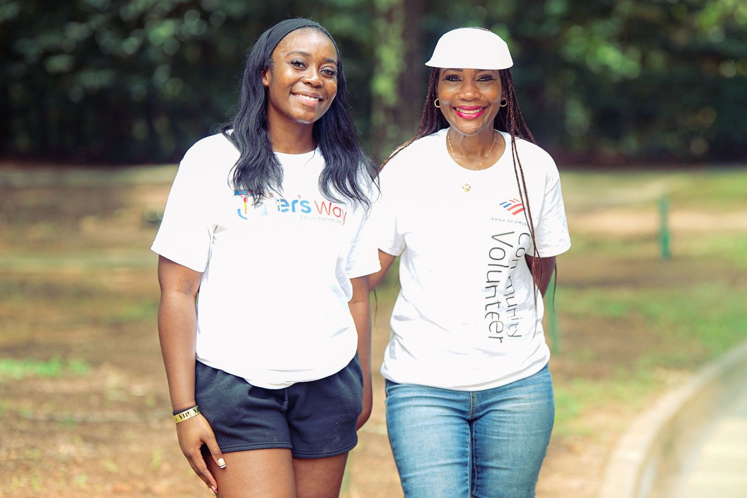 Two women in white volunteer t-shirts smile while walking together outdoors in a park setting.