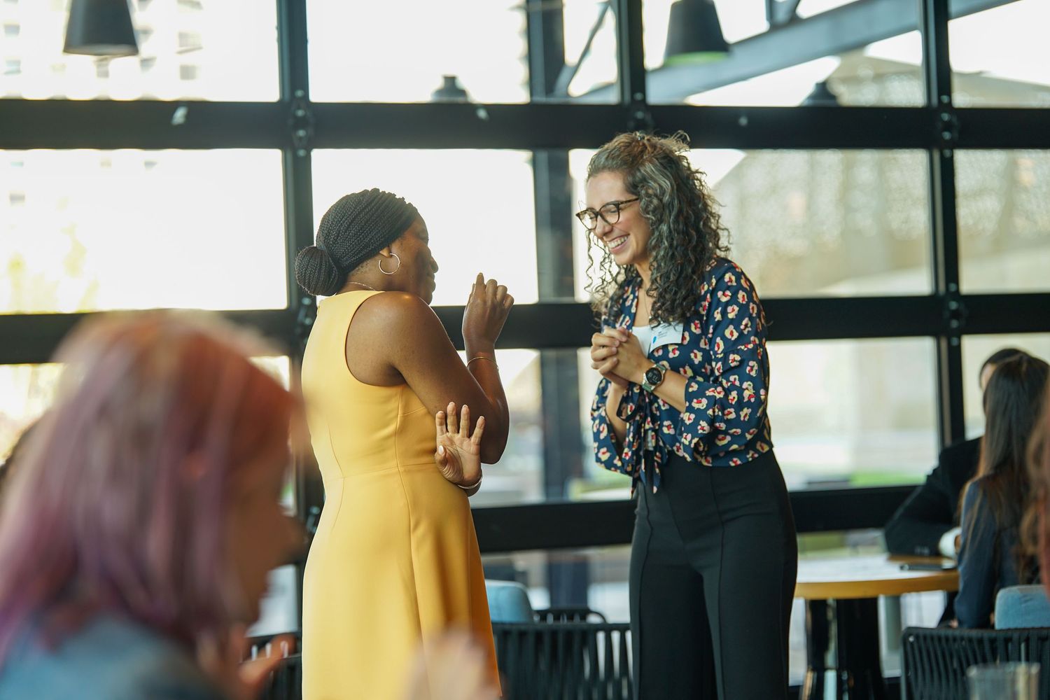 Two women laughing and networking at a professional event in a bright modern venue.