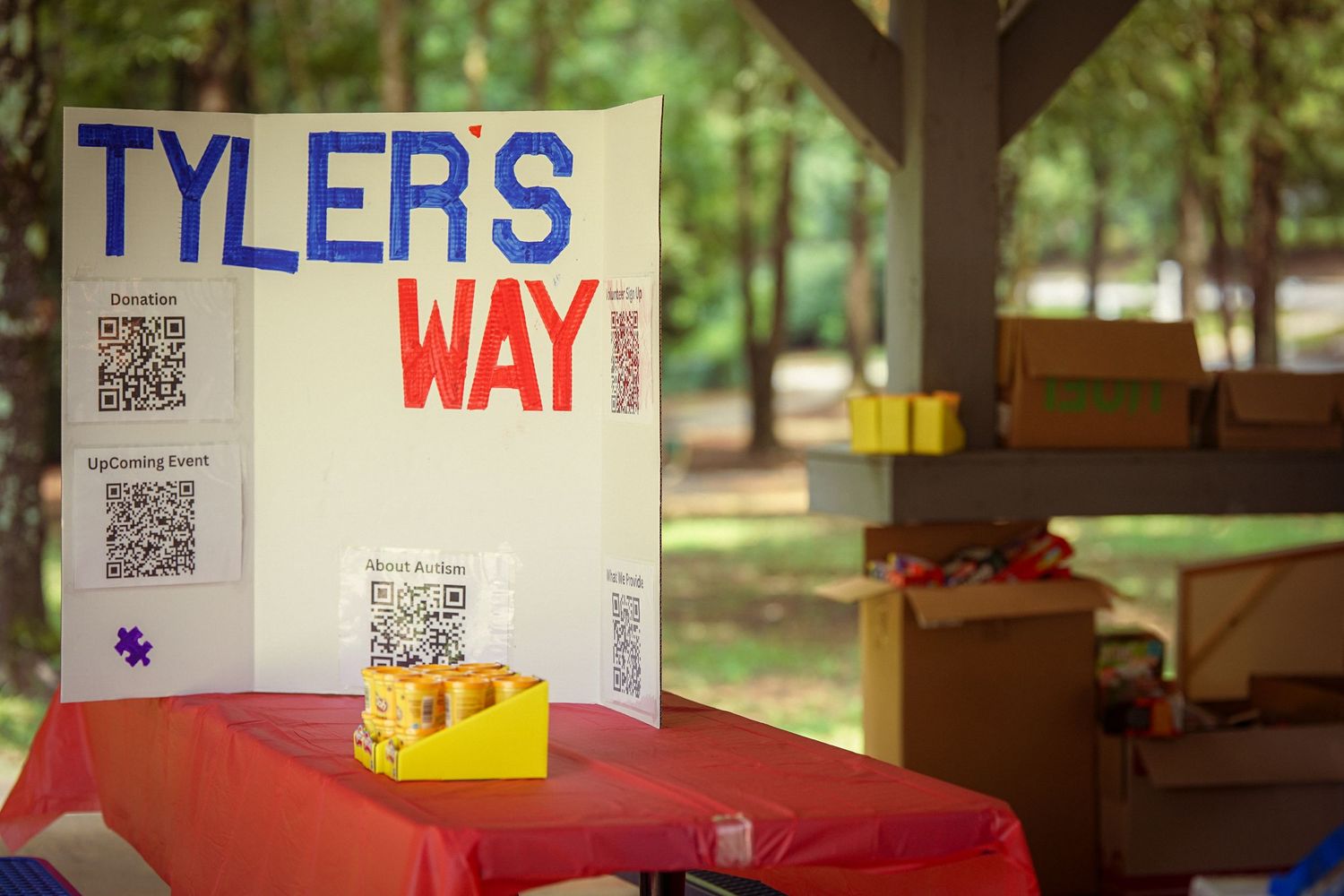 Tyler's Way charity event booth with QR codes and donation sign displayed under a pavilion outdoors.