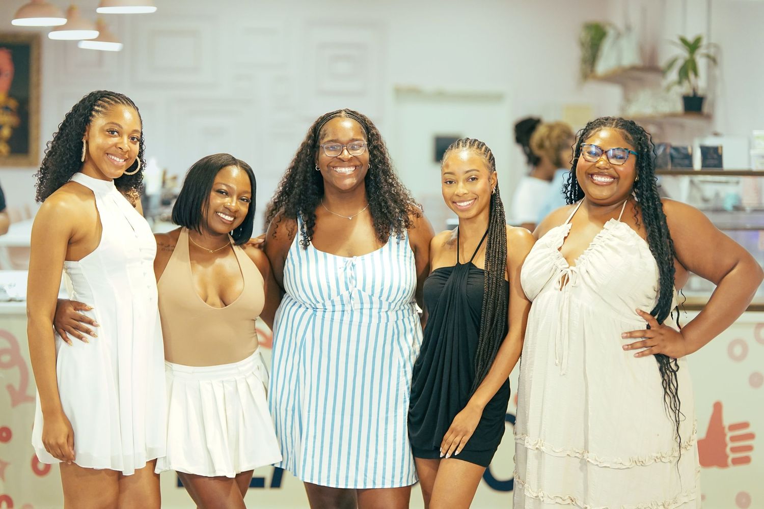 Five young women smiling and posing together indoors, wearing summer outfits at a casual social event.