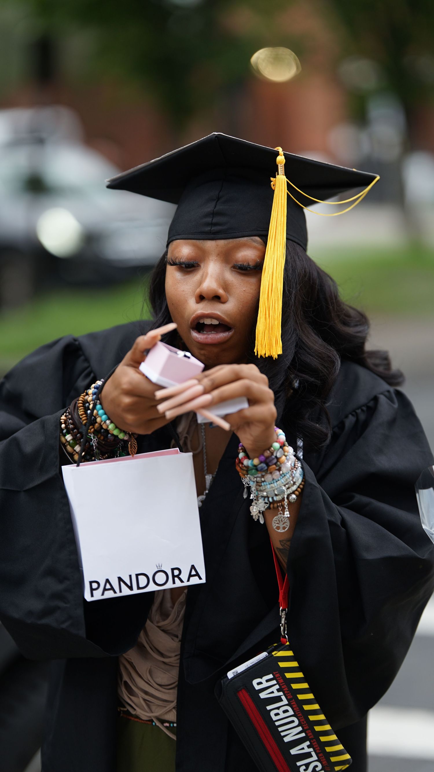Graduate in academic dress poses expressively with Pandora sign outdoors.
